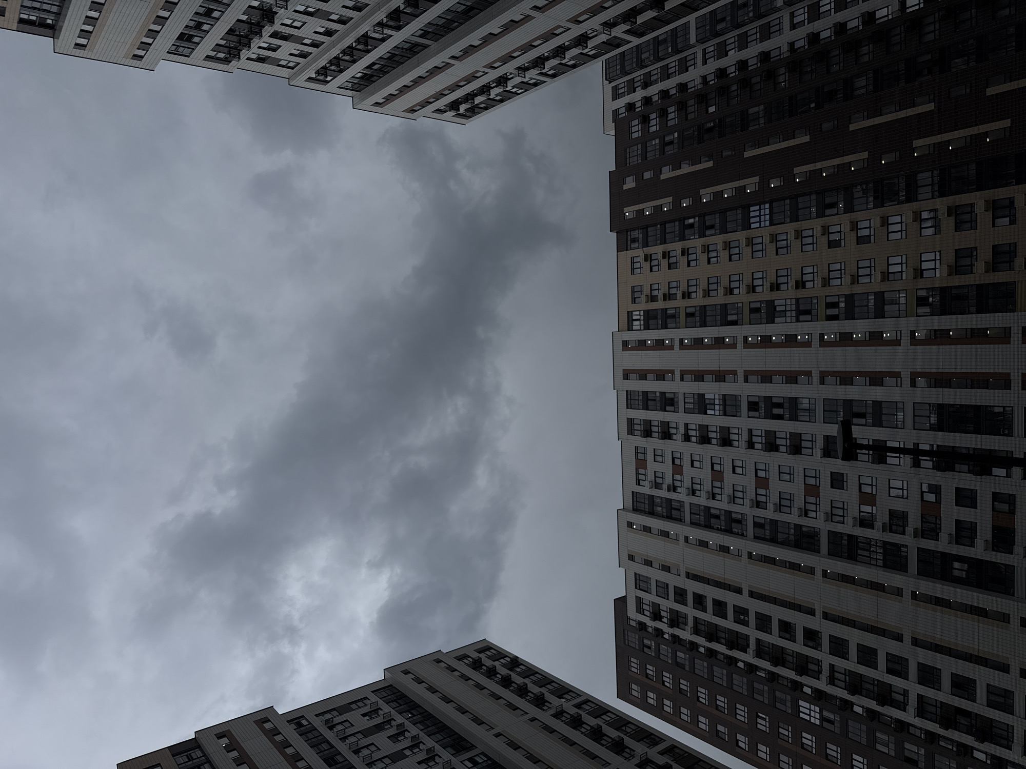 A low-angle shot captures four modern apartment buildings encircling a view of a cloudy, overcast sky. The buildings are predominantly white and gray, with rows of rectangular windows arranged in a grid pattern. One building features dark brown sections. The sky fills the majority of the frame, its gray tones emphasizing a somber mood. The buildings appear located in a densely populated urban area, specifically Lyubertsy, Russia, as indicated by the building style and setting. No people are visible in the scene. The architecture and overcast sky define the environment.