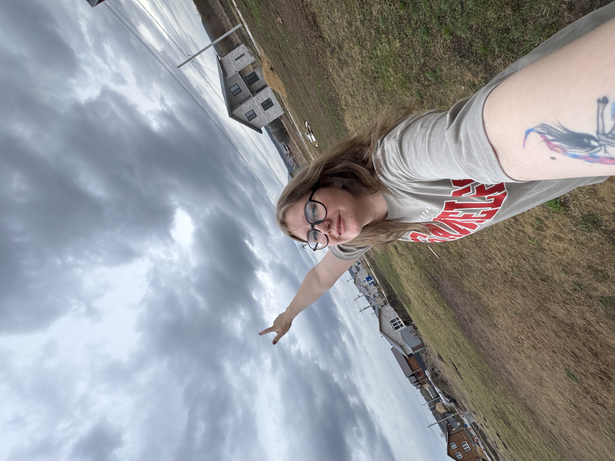 A young woman is featured in a selfie, captured outdoors. She has long, light brown hair and wears black-rimmed glasses. Her right arm is extended towards the camera, with her hand showing a peace sign. She is wearing a light grey t-shirt with "TEXAS TECH" printed in red on the front. A vibrant, colorful tattoo is visible on her upper left arm.
The background reveals a semi-rural setting under a heavily overcast sky. The ground consists of dry, light brown grass and bare earth. In the distance, several houses are visible, some appearing unfinished with grey block walls and dark window openings. Utility poles and power lines extend across the landscape. The sky is filled with dense, grey clouds, indicating an overcast day. The scene suggests a daytime setting in Gornyi (Dvurechenskoe S/P), Russia. The woman appears relaxed, making a peace gesture.