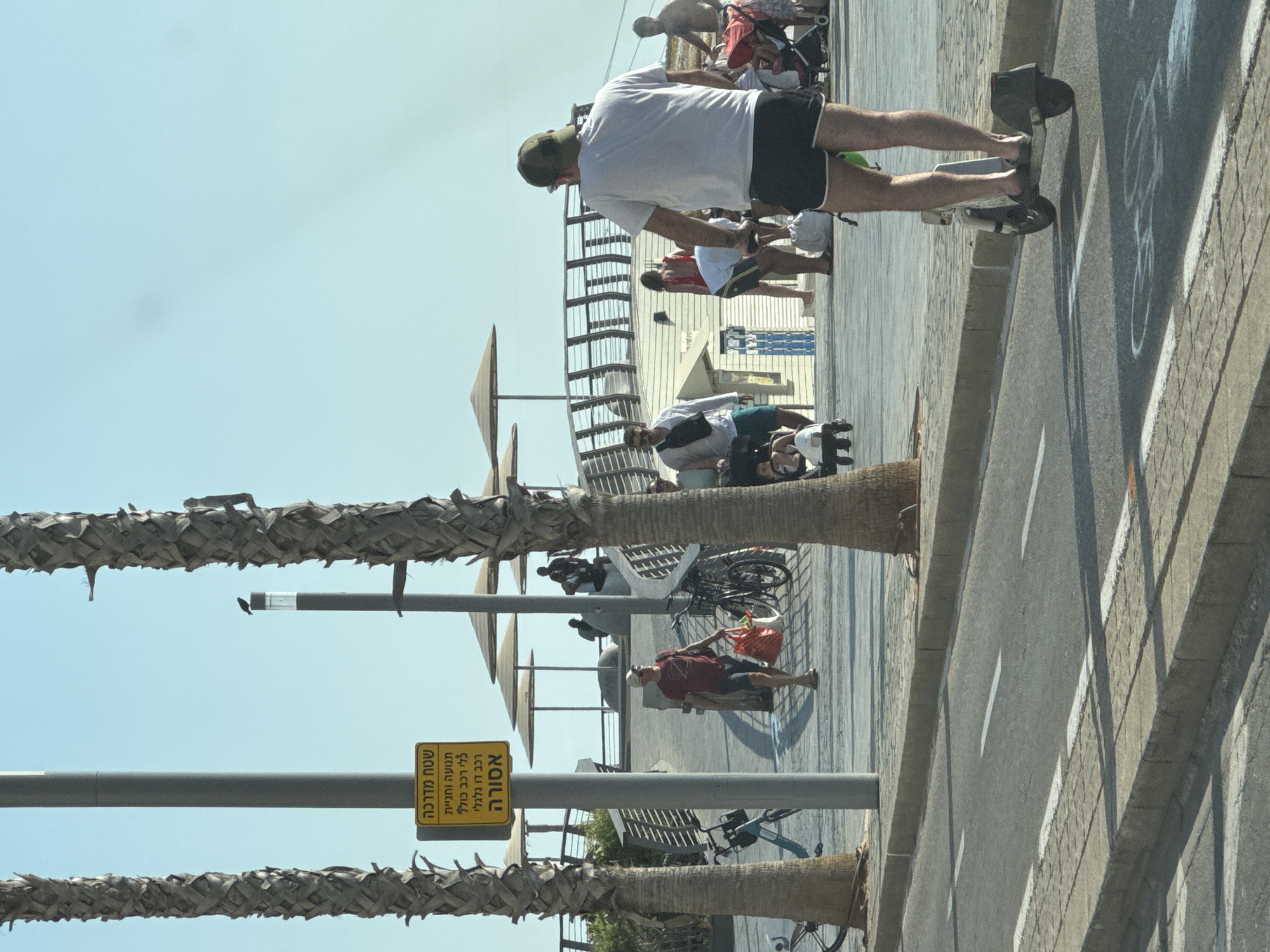 The image captures a scene in Tel Aviv, Israel, likely on a boardwalk or promenade. In the foreground, a man wearing a white t-shirt, dark shorts, and a green cap rides an electric scooter along a paved road. He is facing away from the camera. Behind him, a paved walkway is visible with palm trees flanking the left side of the image. Further in the background, several people are walking and interacting. Some are pushing strollers, and a man in a red shirt is walking with shopping bags. Beach umbrellas are visible in the mid-ground. A yellow sign with Hebrew text is mounted on a pole between two palm trees, indicating a regulation or prohibition. The sky is clear and blue. The overall impression is of a sunny day at a popular public space.