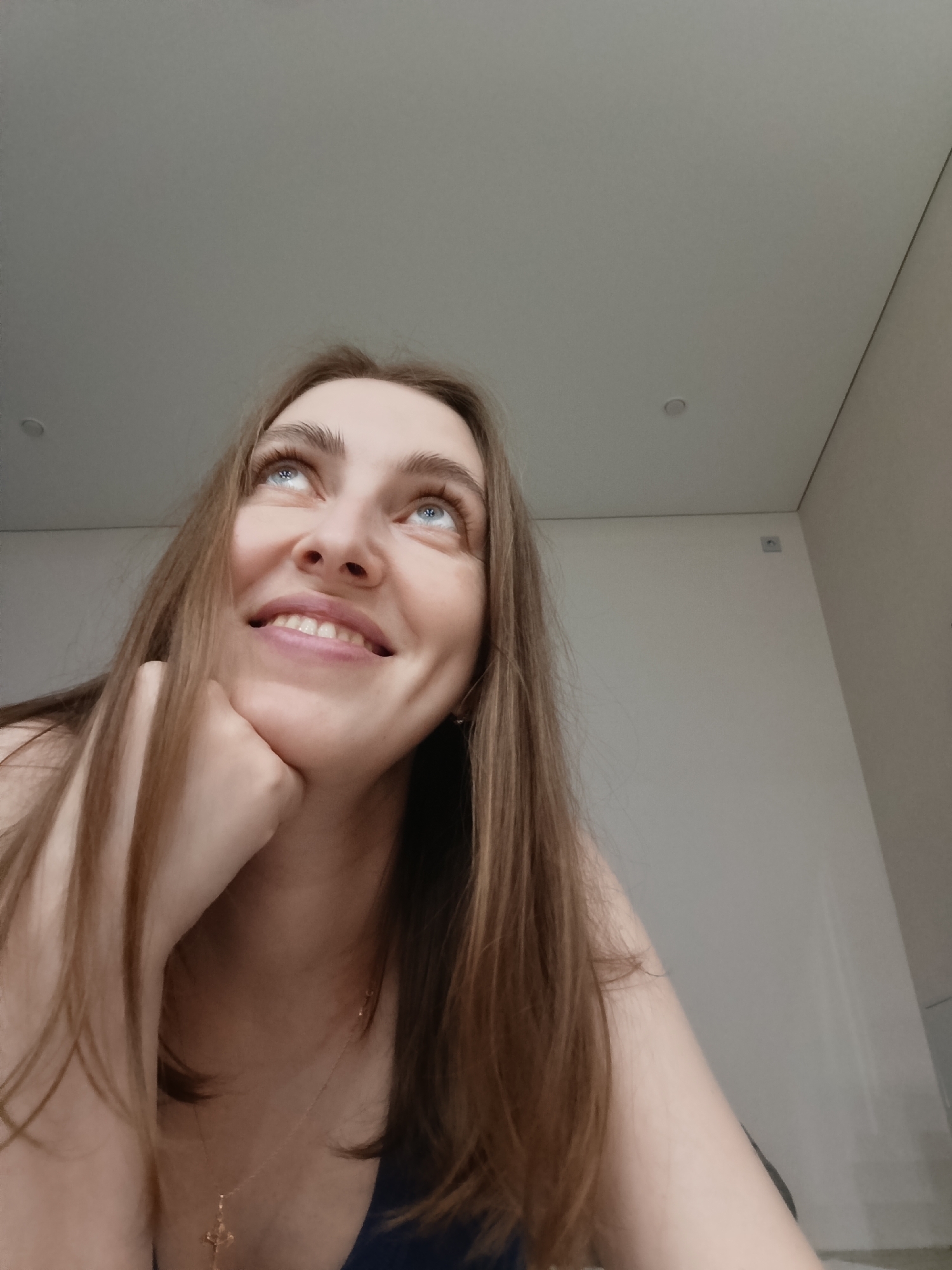 A woman is looking upwards in a room. The woman has light skin, blue eyes, and long brown hair. She is smiling, and her right cheek rests on her right hand. She wears a delicate gold necklace with a cross pendant. The background consists of a white ceiling with a recessed light fixture and a white wall with a molding. I cannot determine the location in the city of Chaikovsky, Russia from this image.