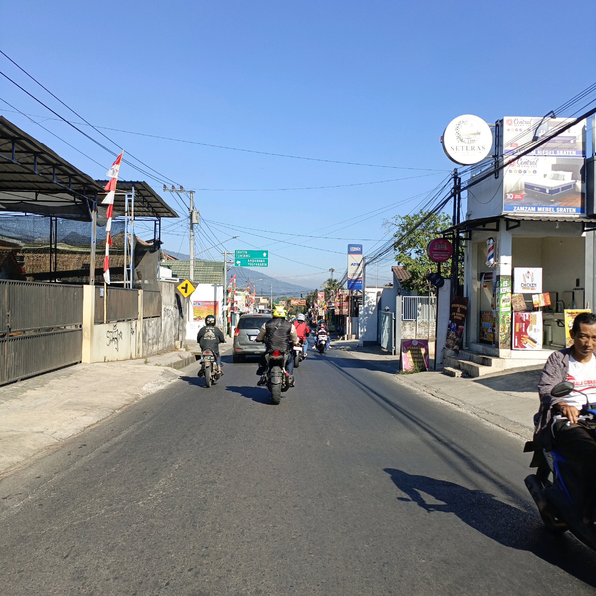 The image depicts a street scene in Sraten, Indonesia, under a clear blue sky. The perspective is from the road, looking forward. A paved road dominates the foreground, with a sidewalk to the left. Several motorcycles are present on the road. In the middle distance, a silver car is visible, along with a green directional sign indicating routes to "SRATEN TUNTANG" and "AMBARAWA YOGYAKARTA". Buildings line the right side of the street, including one with a "BRI ATM" sign and another with advertisements for "ZAMZAM MEBEL SRATEN" featuring mattresses. There are also various smaller shops and businesses. A man is riding a motorcycle on the right side of the road in the foreground, casting a shadow. On the left side of the road, a building with a dark-covered roof is visible, with Indonesian flags displayed. The overall impression is of a typical urban street with ongoing traffic and commercial activity.
