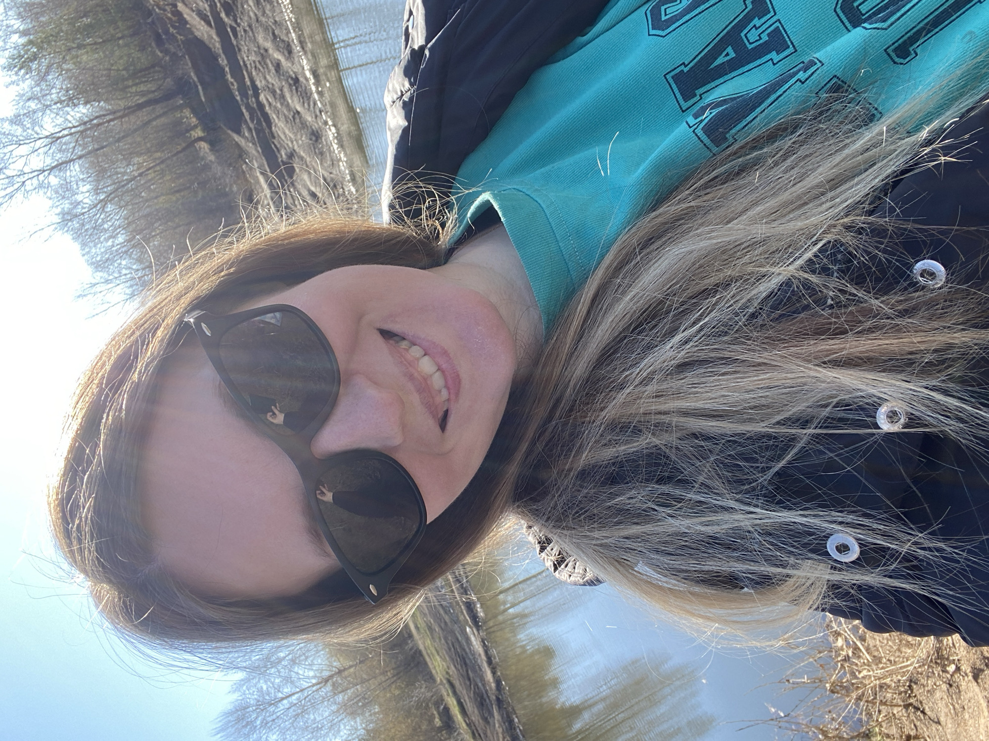 A woman in a black jacket and teal sweatshirt is taking a selfie. She is wearing sunglasses that reflect a hand gesture, possibly a peace sign. The background appears to be a natural environment, potentially near a body of water and trees. Given the request for a description related to Chaikovsky, Russia, it could be presumed this photo was taken in a park or natural area within or near that city. The weather is sunny. The photo is taken at an unusual angle.