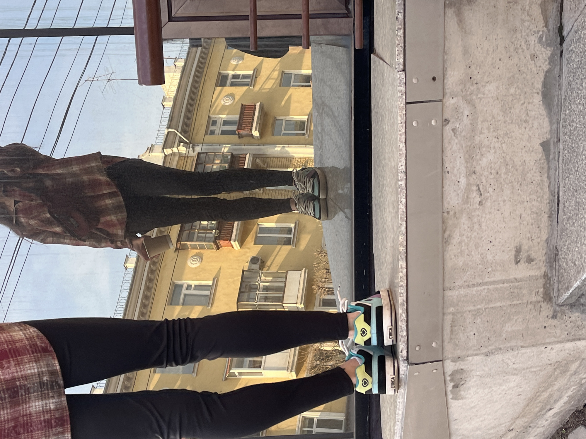 The image captures a reflection in a window. In the reflection, there is a person's lower body and legs, clad in black leggings and a red and black plaid shirt. The person is wearing two different pairs of sneakers. One pair appears to be older, with a muted color palette, while the other pair is brightly colored in teal, lime green, and black with white laces, and labeled "CIRCA" on the side. The window reflects a yellow building with multiple windows and architectural details. The immediate foreground shows a gray concrete or stone surface.