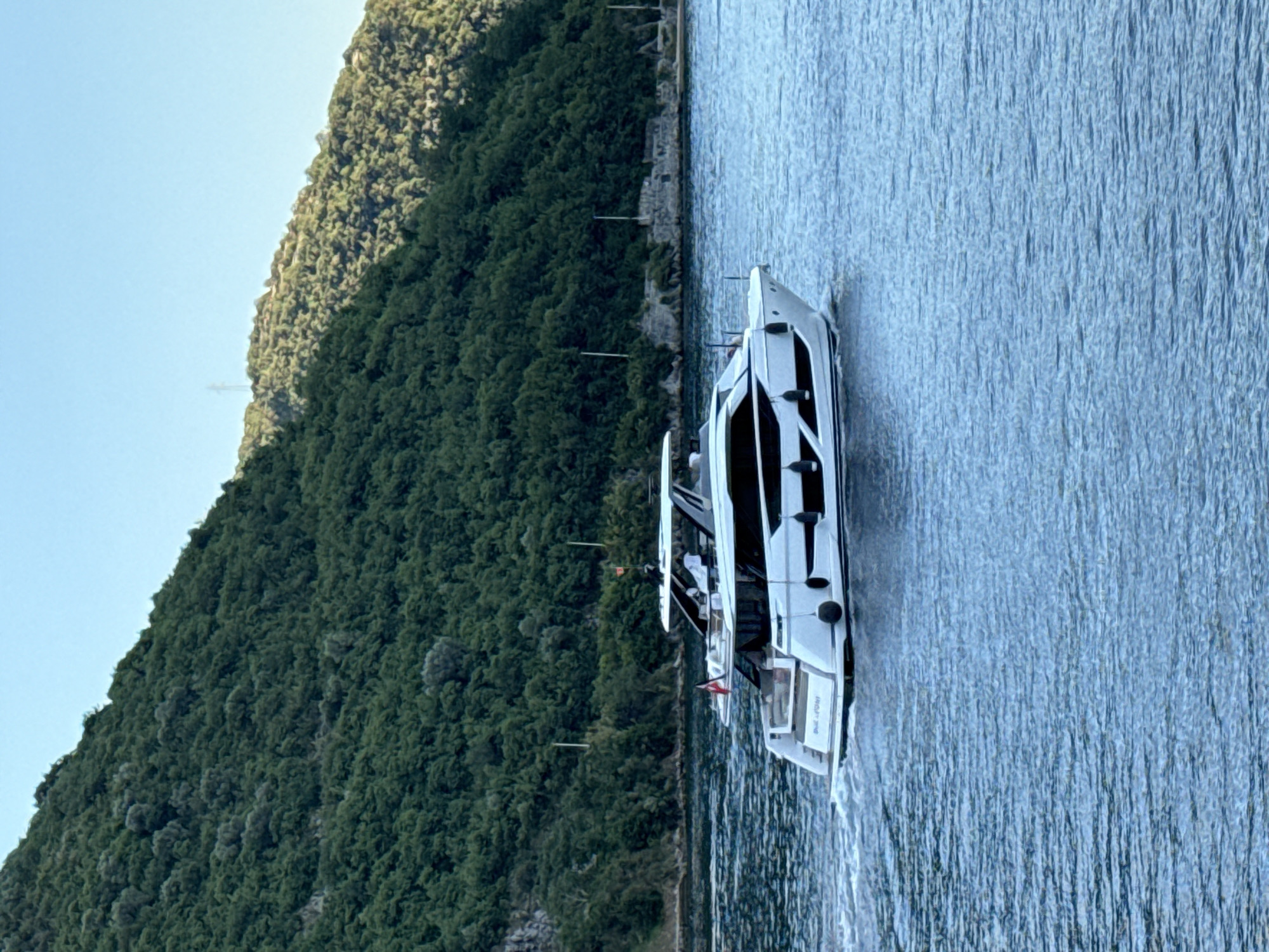 In the city of Đurići, Montenegro, a sleek, white motor yacht is cruising on the calm, blue water. The yacht features dark trim accents and a multi-level design with visible windows and railings. A small flag is flown from the stern. In the background, a dense, green, tree-covered hillside rises steeply from the water's edge. The sky above is a clear, pale blue, indicating a bright, sunny day. The scene captures a moment of leisure and natural beauty.