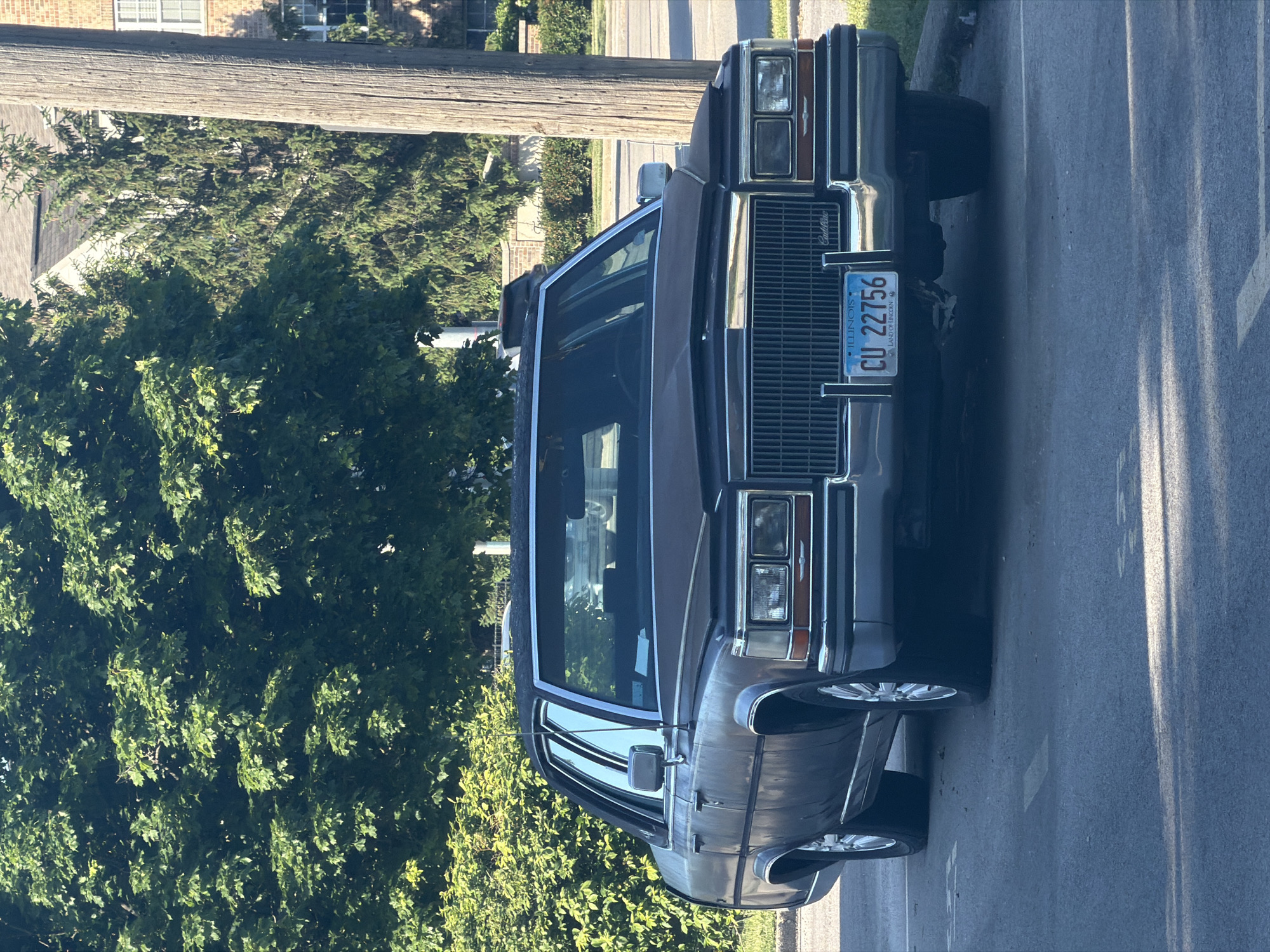 The image shows a dark-colored Cadillac Brougham parked on a paved road or street in Morton Grove, Illinois. The car is facing the viewer. The Illinois license plate "CU 22756" is visible on the front of the car. A wooden utility pole is situated nearby. Green foliage surrounds the car, and there appears to be a building in the background.