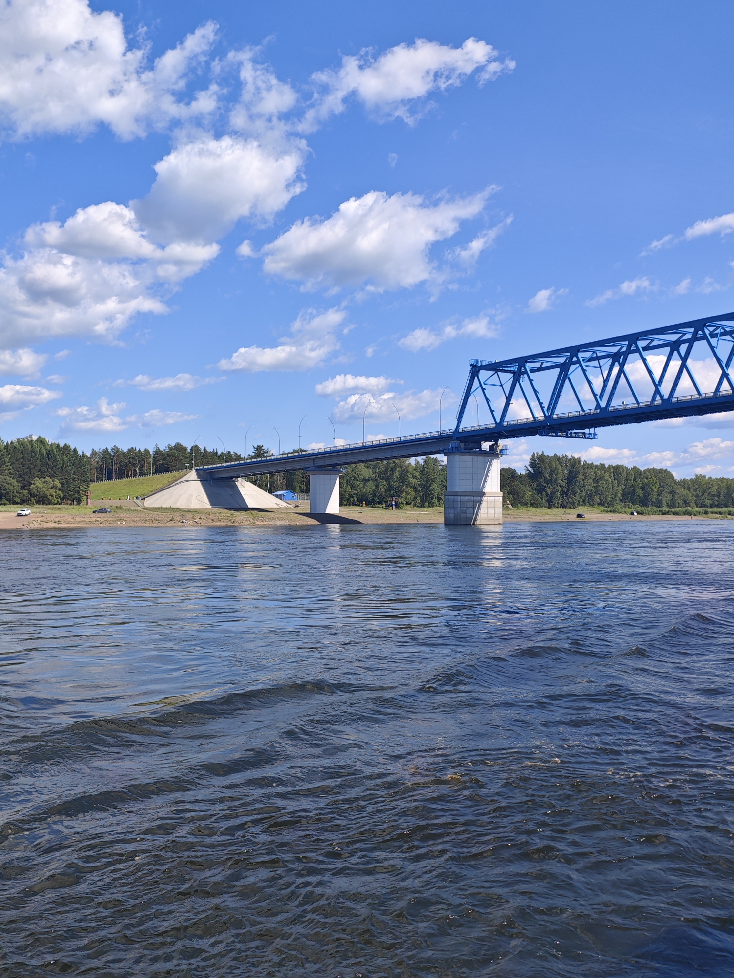 A wide, blue truss bridge spans a dark, rippling river under a bright blue sky dotted with fluffy white clouds. The bridge has a complex latticework structure. On the left bank, a grassy slope rises towards the bridge, with scattered trees and a few distant figures and vehicles visible. The right bank is lined with dense green trees. The foreground is dominated by the textured surface of the river, with waves creating patterns of light and shadow. The scene appears to be a scenic view of the bridge in Abalakovo, Russia.