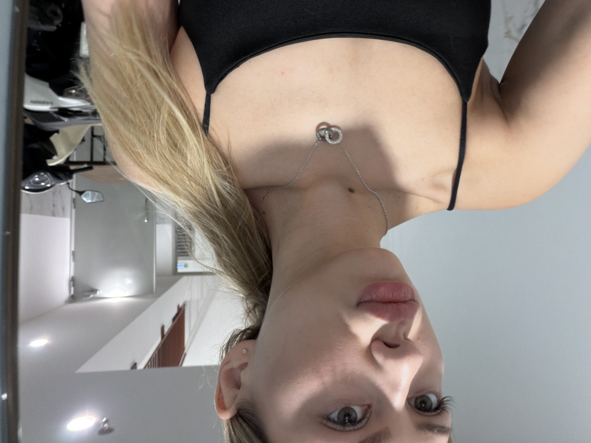 This is a selfie taken indoors of a young woman with blonde hair. She is wearing a black tank top and a silver necklace with two interlocking rings. Her head is tilted, and she is looking directly at the camera with a neutral expression. Her lips are slightly parted, and she is wearing subtle makeup. A small stud earring is visible in her left ear.
The background suggests an indoor environment with a modern aesthetic. The walls are light-colored, and there are several recessed lights on the ceiling, indicating an artificial light source. To the left of the woman, there is a glimpse of a hallway or room with what appears to be a staircase with wooden railings and a white wall. There are also some objects that could be part of furniture or decor. The overall lighting is bright and even.
There is no visible text in the image. The time of day is not discernible from the lighting conditions. The location is identified as Đà Nẵng, Vietnam, though no specific landmarks or cues in the image definitively point to this location. The activity is simply posing for a photograph.