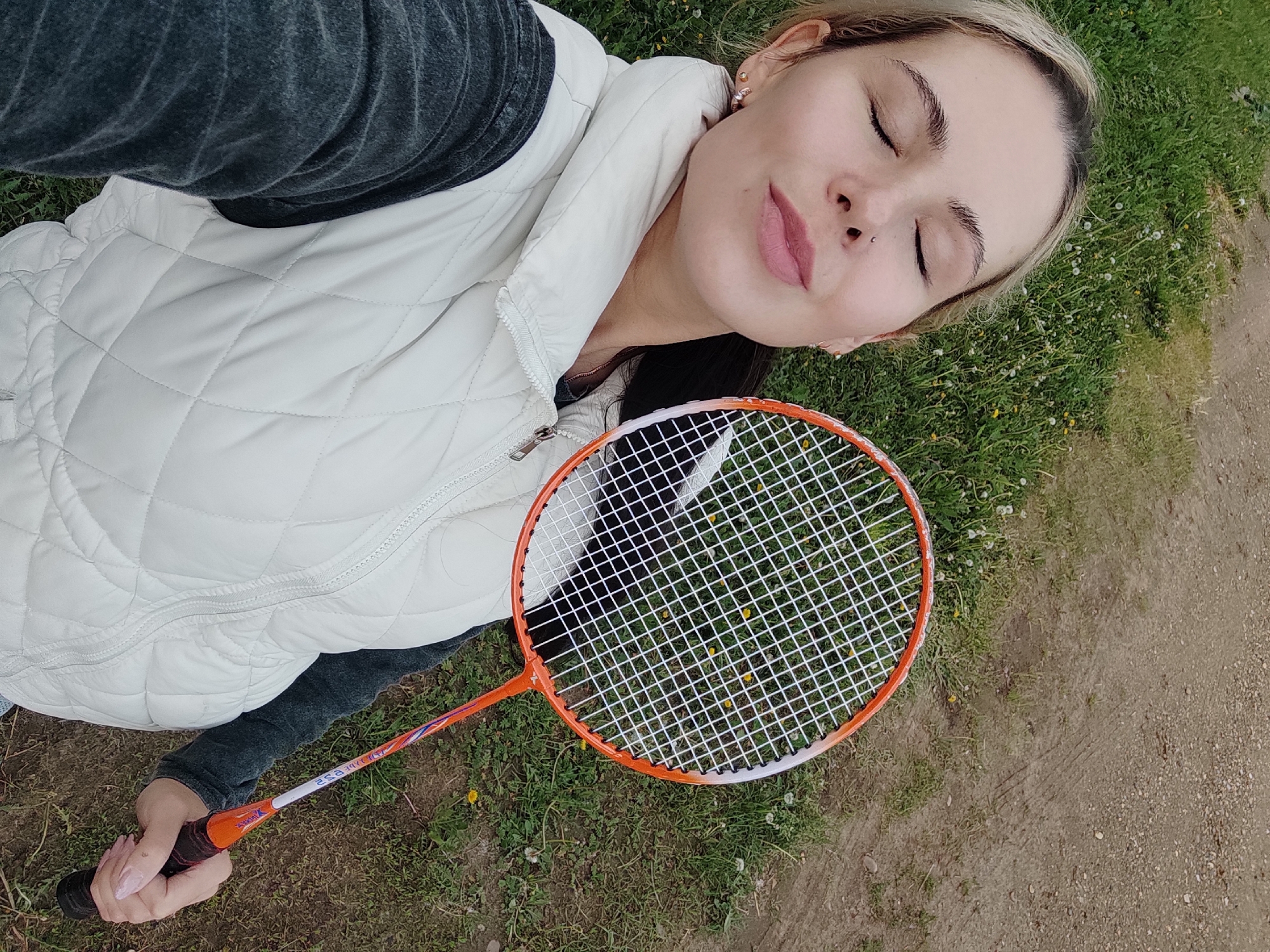 A young woman lies on a grassy surface with her eyes closed. She wears a white quilted vest over a dark gray garment. She is holding a badminton racket with an orange frame. The location appears to be outdoors, with grass and a dirt path visible. The image was likely taken in Ольховка, Russia.