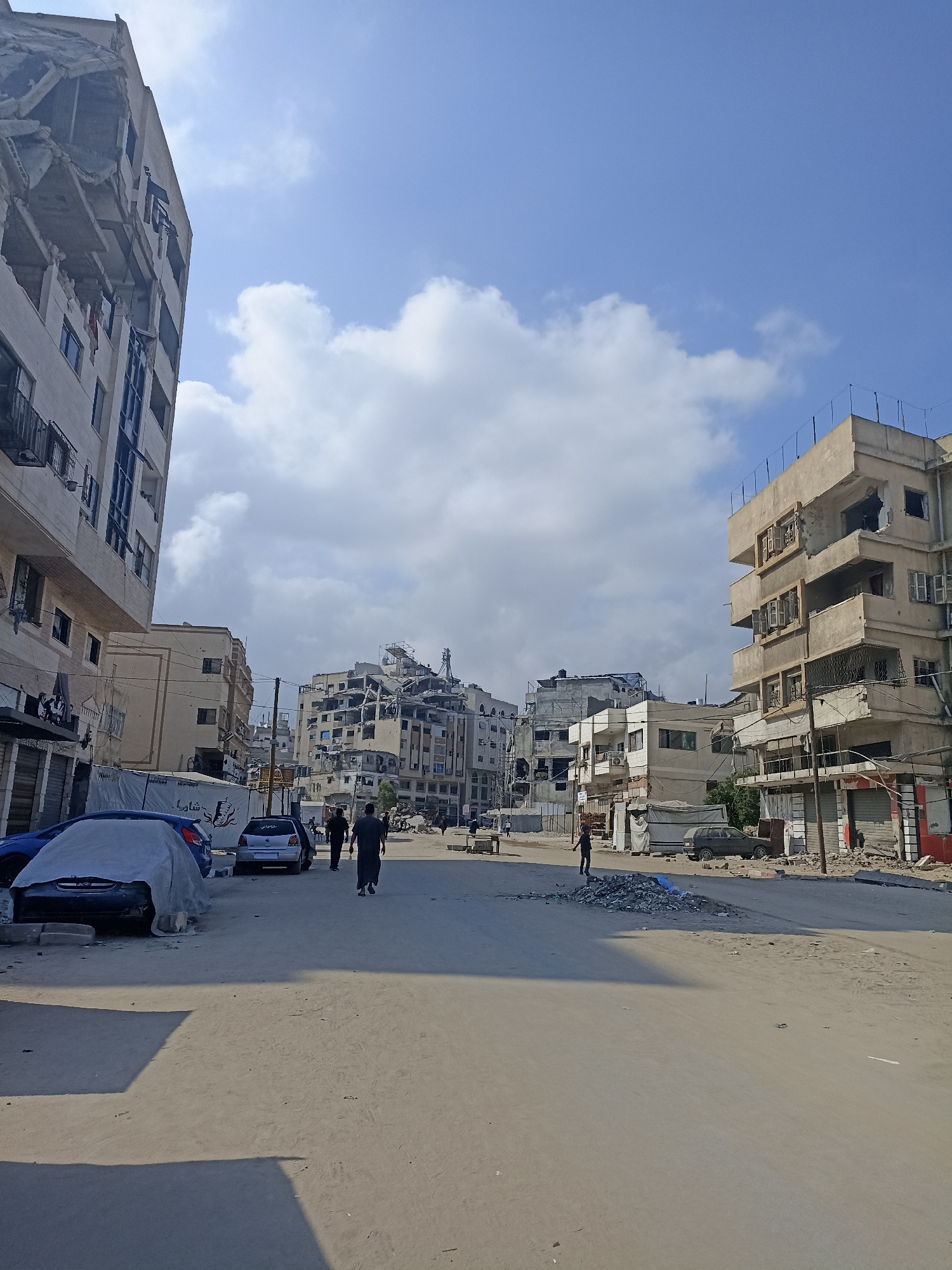 The image depicts a street scene in Gaza City, Palestinian Territories, with several damaged buildings on either side of a paved road. The buildings show signs of destruction, with some upper floors collapsed or heavily damaged, exposing internal structures and rebar. One building on the left has its top floors severely damaged, appearing partially caved in. On the right, a multi-story building exhibits shattered windows and significant structural damage to its facade. The street itself is dusty and appears to be undergoing some form of cleanup or reconstruction, with piles of rubble visible on the roadside. A few cars are present; one is covered with a tarp on the left, and a silver sedan is parked further down the street on the right. Several individuals are walking along the street. Two men are walking in the middle ground, one wearing dark clothing and the other in lighter attire. Another person, possibly a child, is standing near a pile of rubble on the right side of the road. The sky is blue with scattered clouds. The overall scene conveys an atmosphere of post-conflict devastation and ongoing efforts to deal with the aftermath.
