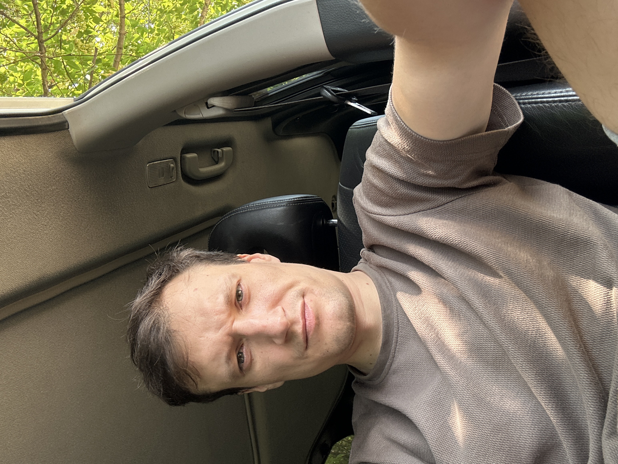 A man is seated in the driver's seat of a car, his face partially obscured by a selfie. He is wearing a light brown, textured t-shirt. Sunlight filters through the trees outside the car, casting shadows on his face and shirt. The interior of the car is visible, including the headrest of the passenger seat and part of the dashboard. The image appears to have been taken during the day. There is no discernible information within the image to indicate the specific location within the city of Makiv, Ukraine.