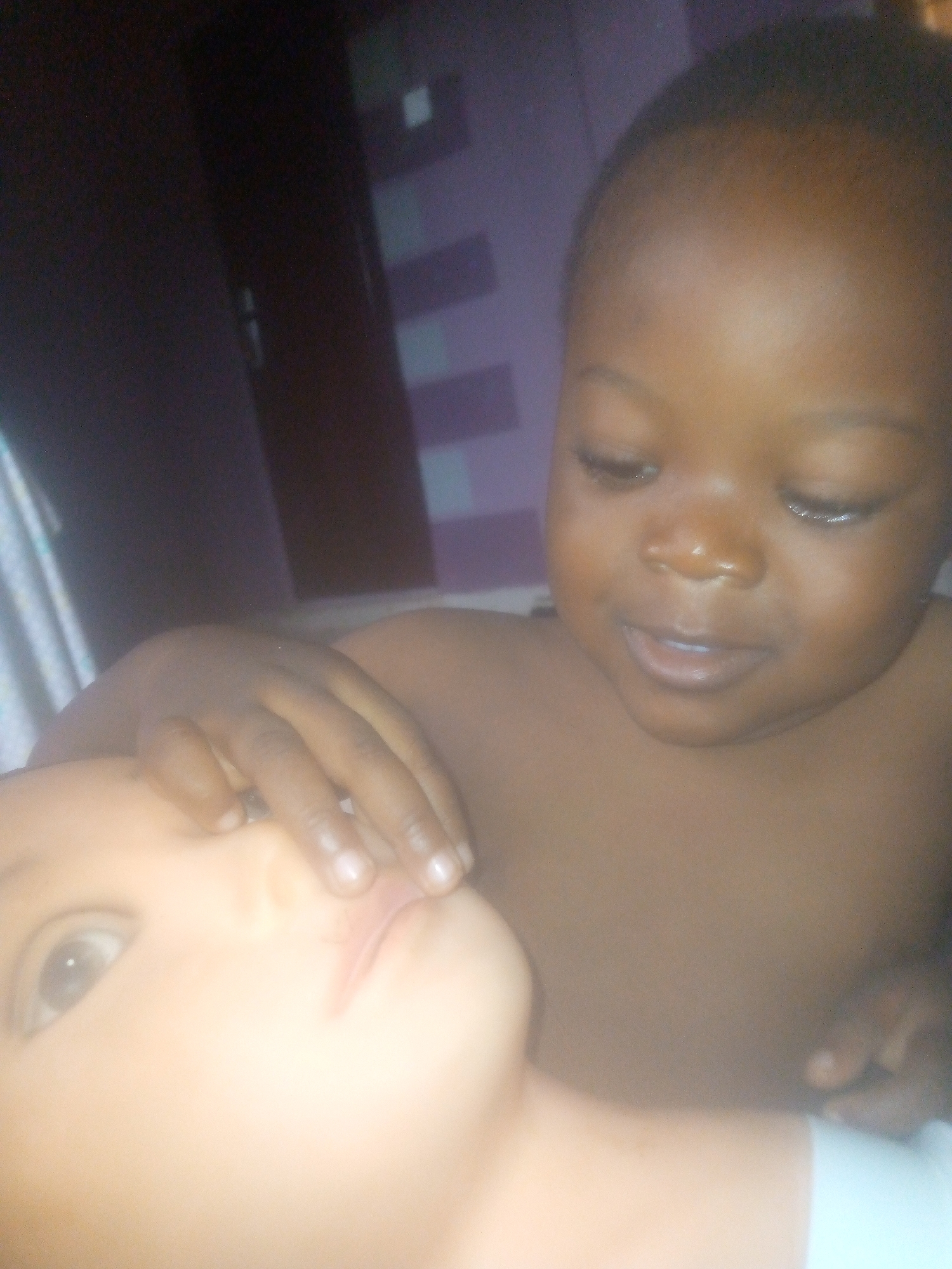 A dark-skinned baby with a smooth, bald head is shown in close-up, looking downward with a slight smile. The baby's right hand rests gently on the face of a pale-skinned doll, which is lying on its back. The background appears to be an interior room, with a dark brown door visible on the left and a striped white and purple design partially visible behind the baby's head. The location is Unknown, Unknown.