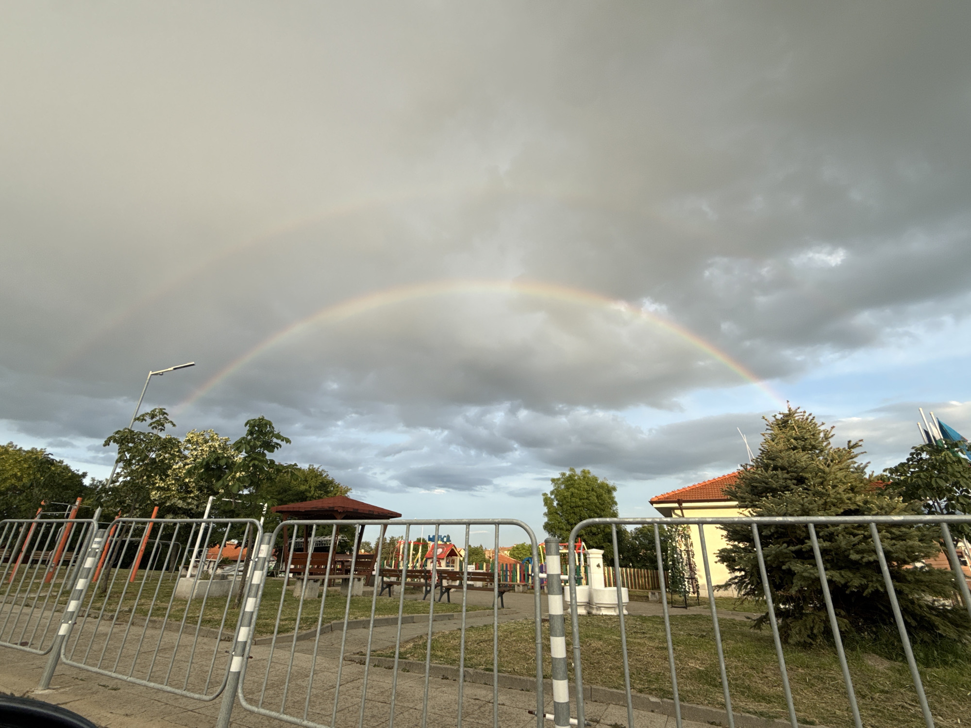 The image shows a park in Ivanyane, Bulgaria, on a cloudy day with a double rainbow visible in the sky. The foreground is occupied by a metal fence. Behind the fence, there is a playground with benches, a gazebo, and children's play equipment. A street lamp is visible to the left, and a large fir tree stands in the middle right of the image, near a building with a red-tiled roof. The sky is filled with dramatic, grey clouds, suggesting a recent or impending rain shower. The overall mood is one of nature's beauty amidst a common outdoor setting. The time of day appears to be late afternoon or early evening, given the lighting. There is no visible text in the image.