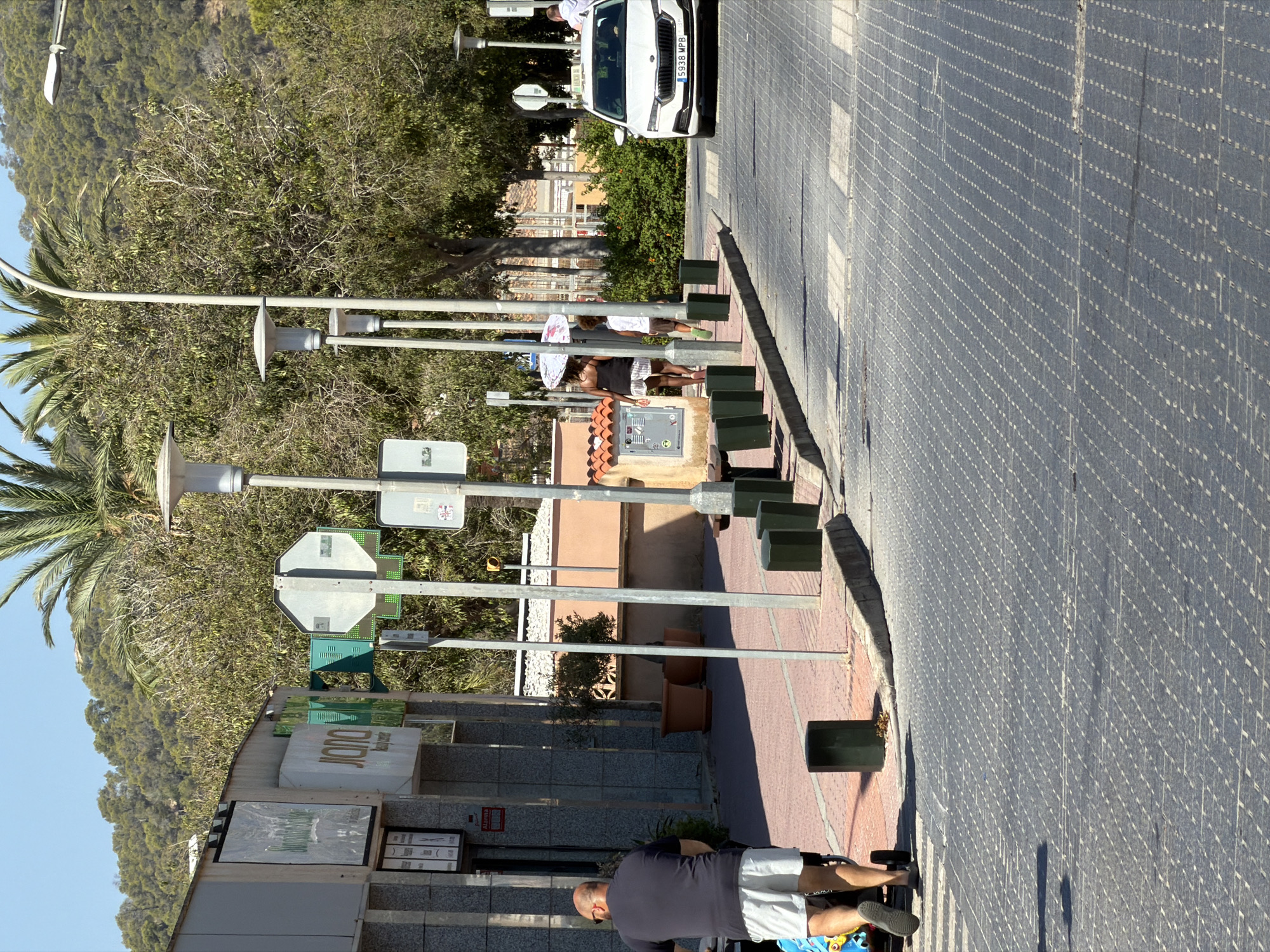 The image shows a street scene in Calvià, Spain, on a sunny day. In the foreground, a man is pushing a stroller along a pedestrian walkway. The walkway is paved with cobblestones and bordered by a curb and a street. A crosswalk is visible on the street. In the background, there are buildings with storefronts, trees, and a hillside covered in vegetation. A white car, possibly a taxi, is driving on the street. Several streetlights and traffic signs are also present. The environment appears to be a commercial or urban area with pedestrian access.
