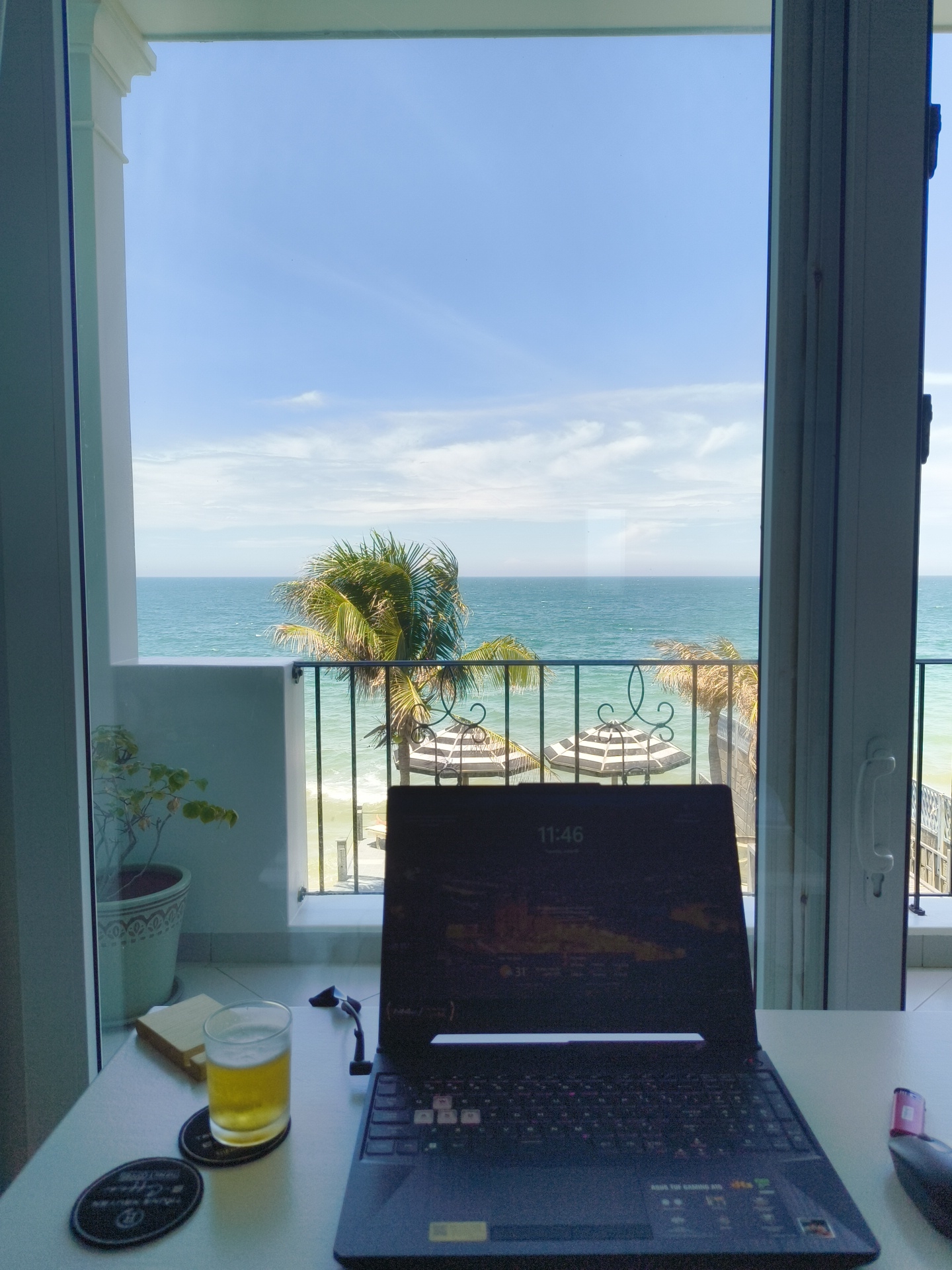 The image depicts a desk area within a room overlooking a balcony and the sea. On the desk, a laptop is open, displaying the time "11:46" and a background image. To the left of the laptop, a glass of beer sits on a coaster. Another coaster is visible to the left of the beer glass. A small potted plant is positioned on a ledge to the left of the desk. Beyond the desk, a glass door opens to a balcony. Through the balcony, a view of the ocean is visible, with a palm tree, two striped umbrellas, and lounge chairs. The scene suggests a relaxed atmosphere, possibly a vacation or remote work setting. The location is identified as Phu Thuy Ward, Vietnam, likely a coastal area. There are no people present in the image.