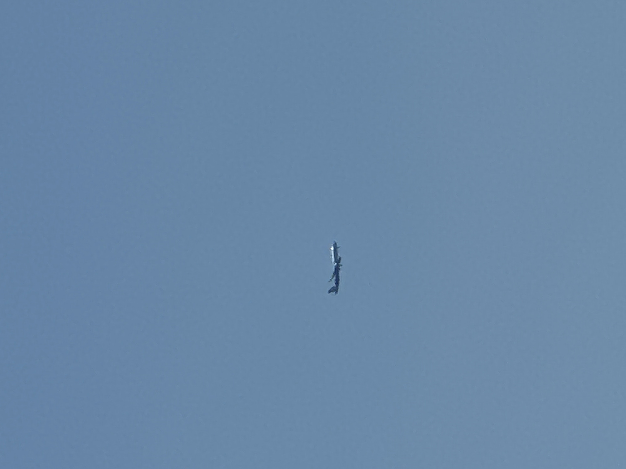 An airplane is seen flying in a clear blue sky. The airplane is a light color with dark accents. Its wings are extended and its engines are visible. The sky is a uniform light blue, indicating daytime and good weather. There are no other objects or people visible in the frame. The image does not provide any information about the city of Boreti, Montenegro.