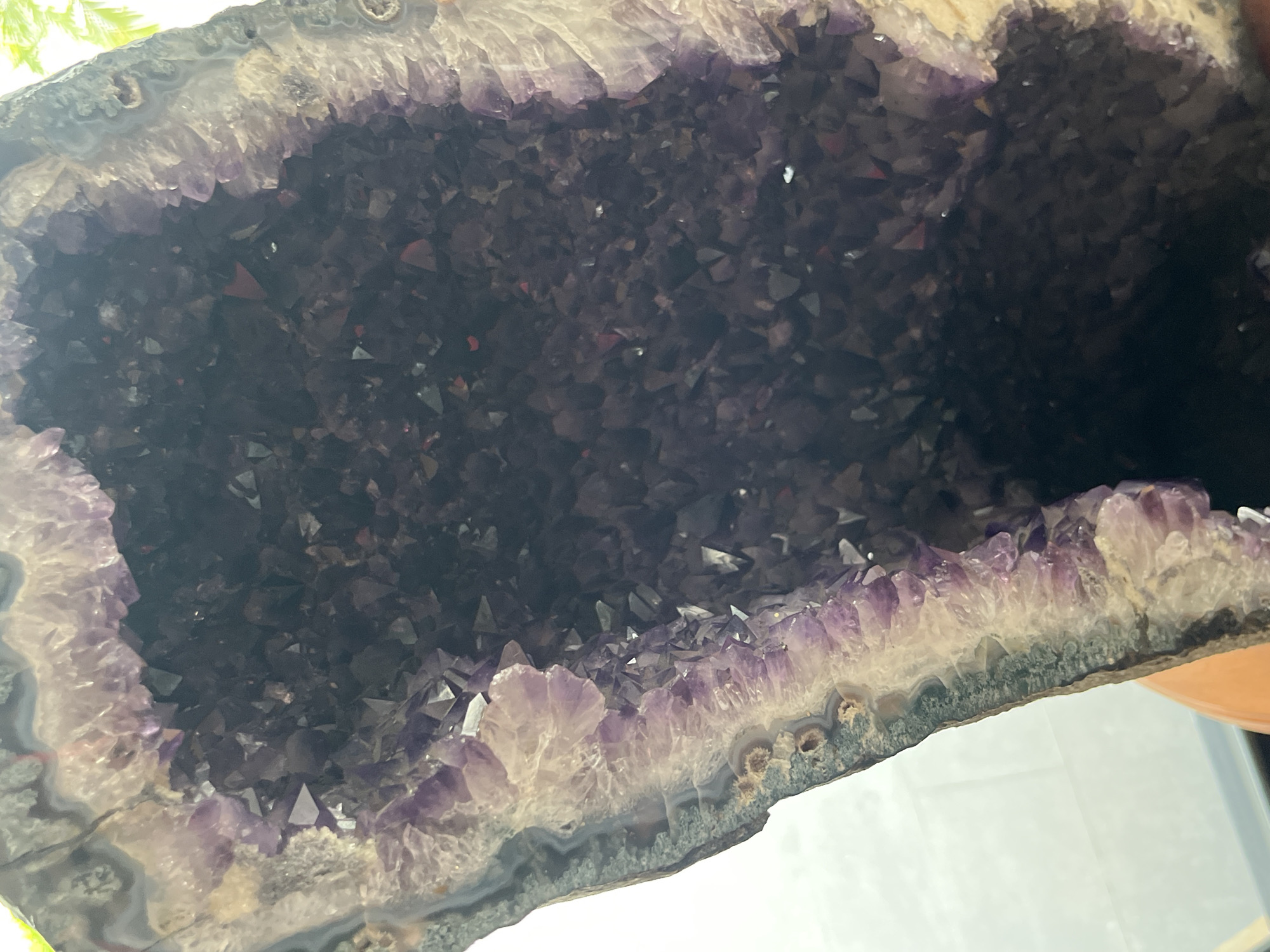 The image shows a close-up of a large amethyst geode. The geode's exterior is gray with white and darker gray banding. Inside, numerous purple amethyst crystals line the interior cavity. The crystals vary in size and shade, with some appearing lighter purple and others much darker. The photograph appears to be taken indoors under natural light. There is no evidence this geode is in Pattaya City, Thailand.