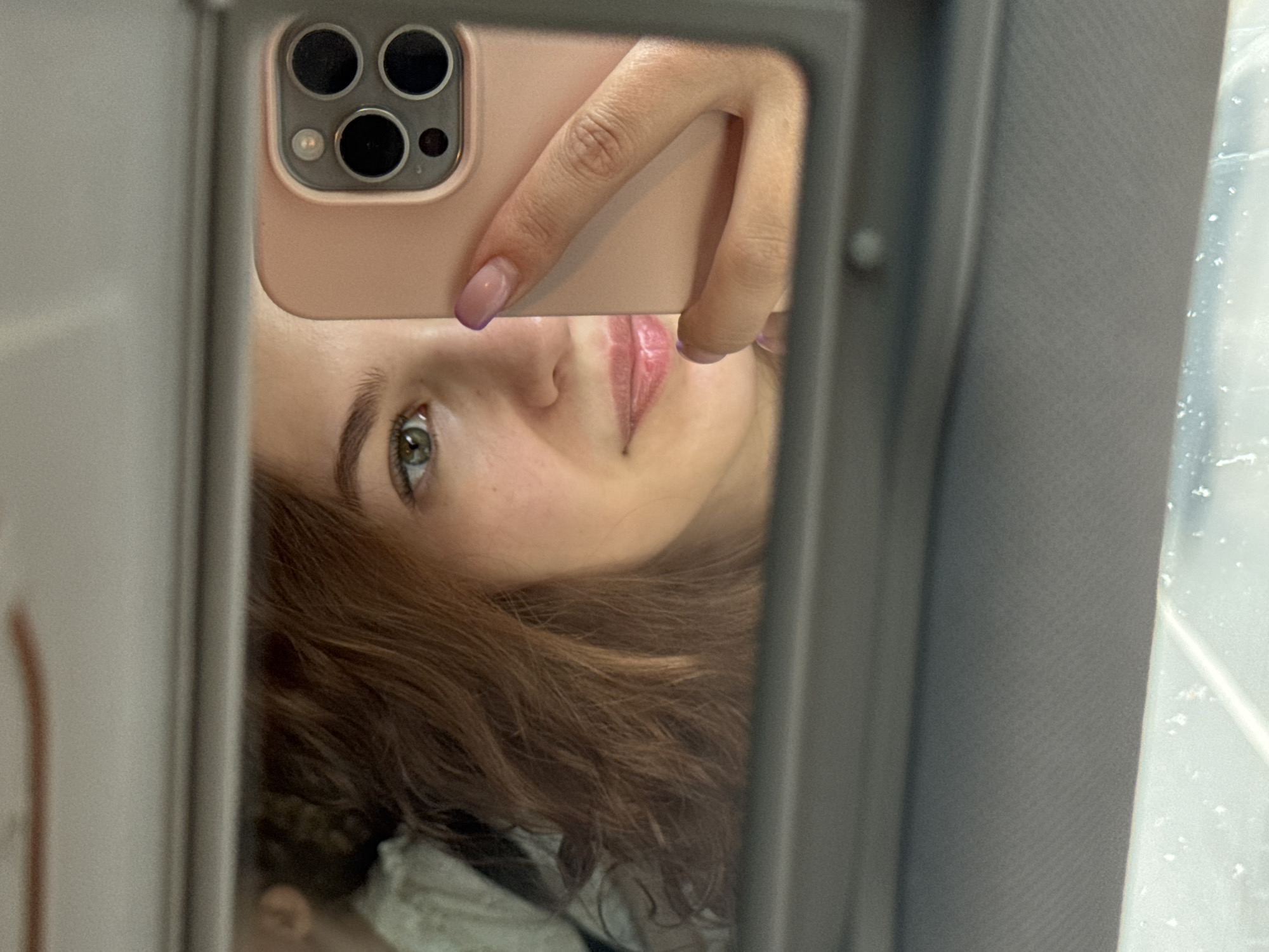 The image is a selfie taken in a mirror, likely in a vehicle. A young woman is holding a smartphone with a light pink case that has three camera lenses. Her face is partially visible, showing one eye, part of her nose, and her lips. She has light brown hair. The mirror is framed by a gray trim, possibly part of a car's visor. In the background, outside of the vehicle, a window shows signs of moisture or condensation. There is no information to suggest this image was taken in the city of Chaikovsky, Russia.
