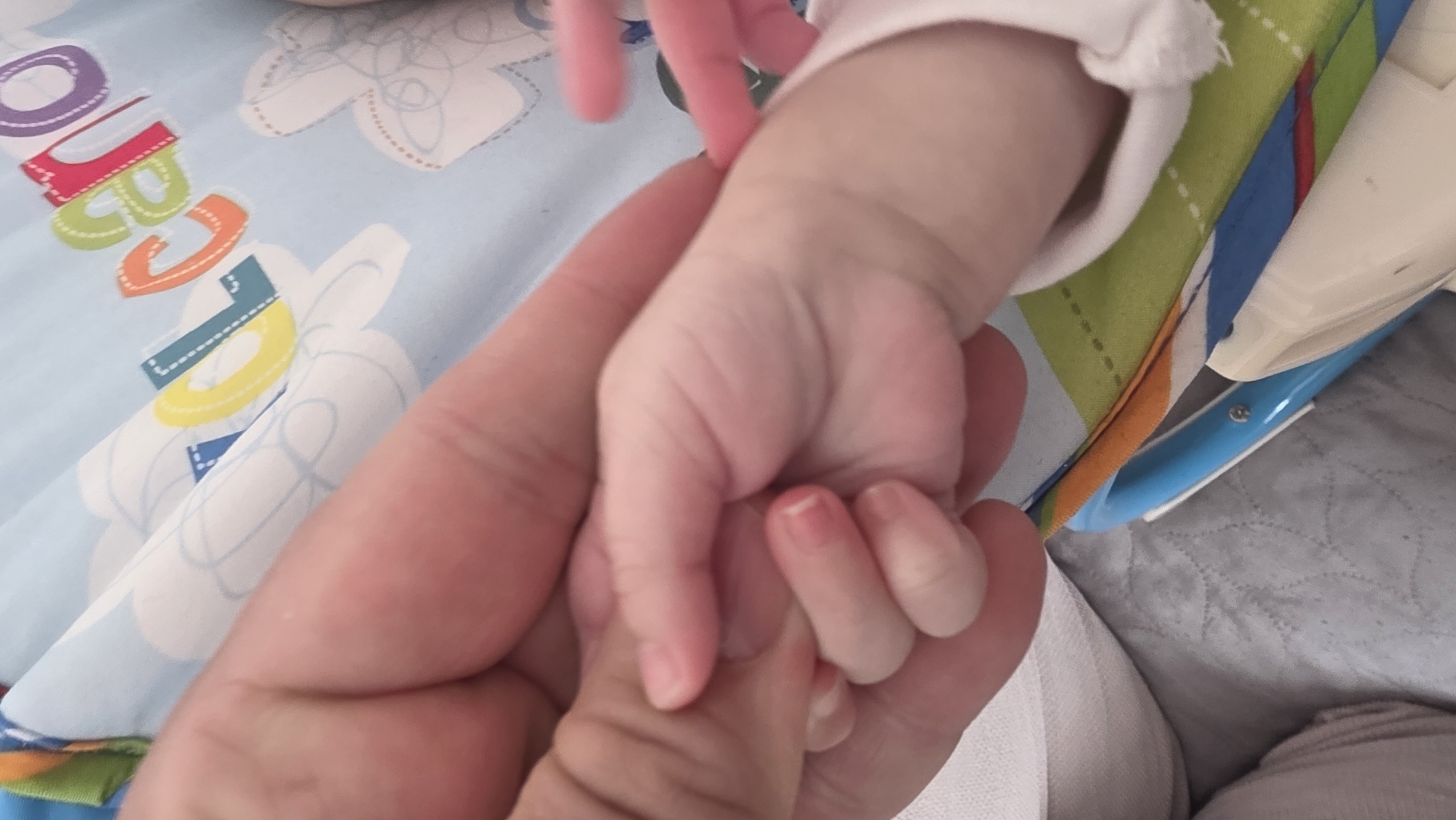 A close-up shot captures a baby's hand gripping an adult's finger. The baby's hand is pink, with tiny fingernails, while the adult's hand has a warm skin tone. The baby is wearing a white sleeve. The background includes a colorful patterned fabric with letters and cloud designs. Part of a highchair is visible with blue and green accents. The location is not discernible in the image and cannot be placed in Blagoevgrad, Bulgaria.