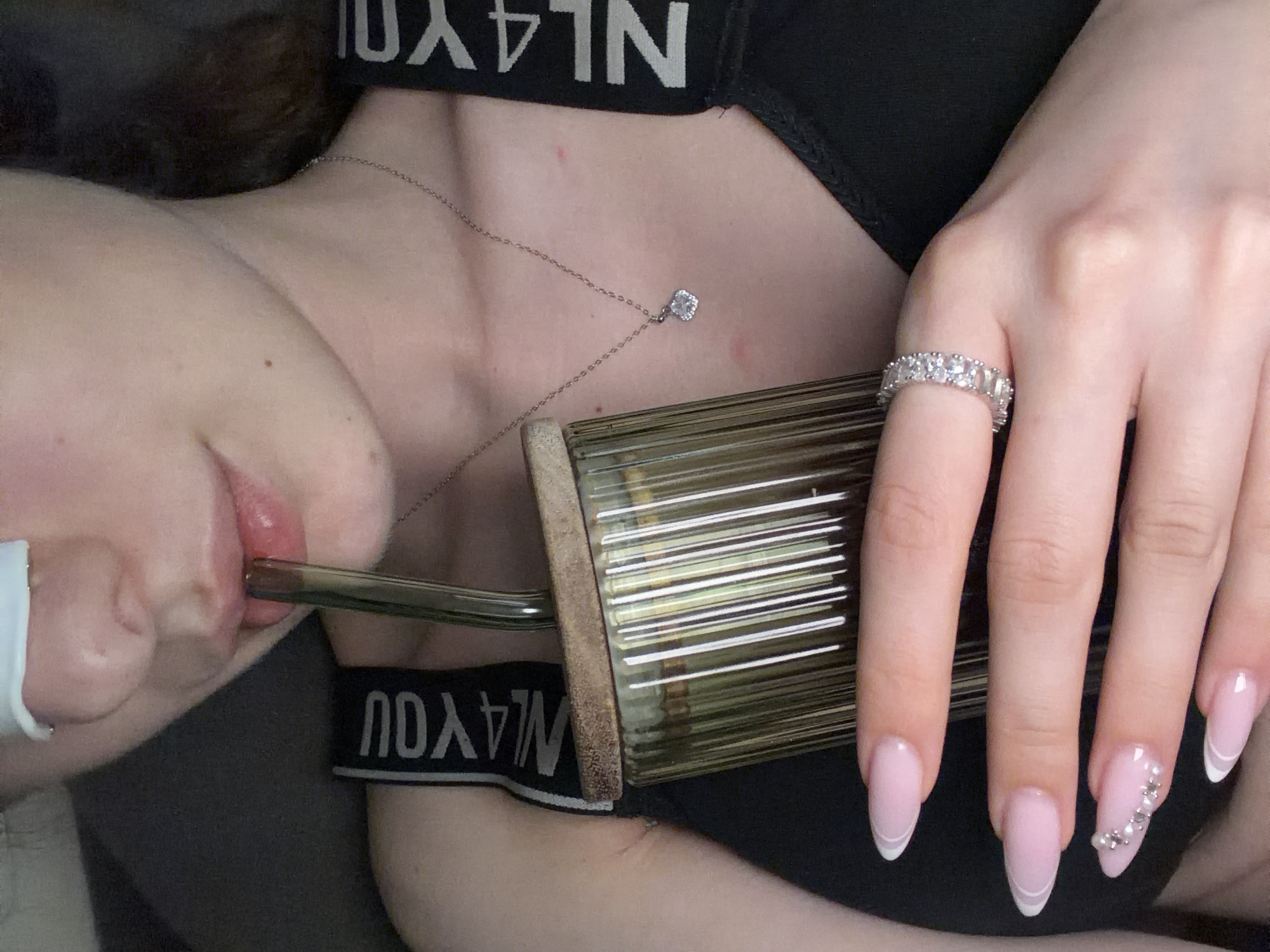 The image provides a close-up, intimate view of a person's upper body, focusing on their chest, neck, lips, and right hand. The person is wearing a black bra or top with a visible elastic band featuring white text. A delicate silver chain necklace with a small, square, sparkling pendant rests on their chest.
The person's right hand is prominent, showcasing neatly manicured nails with a light pink base and white French tips. One nail, likely the ring finger, is additionally decorated with small, silvery beads or gems arranged in a curved design. A sparkling ring, encrusted with multiple clear stones, is worn on one finger.
The primary activity depicted is the person drinking from a transparent, ribbed container through a curved glass straw, which is held to their lips. The container has a somewhat aged or textured rim.
Visible on the black elastic band of the clothing, appearing upside down and mirrored, are the letters "N L 4 Y O U". The lighting suggests an indoor setting, and no specific location or time of day can be determined from the cropped view. The image highlights personal accessories and a casual moment.