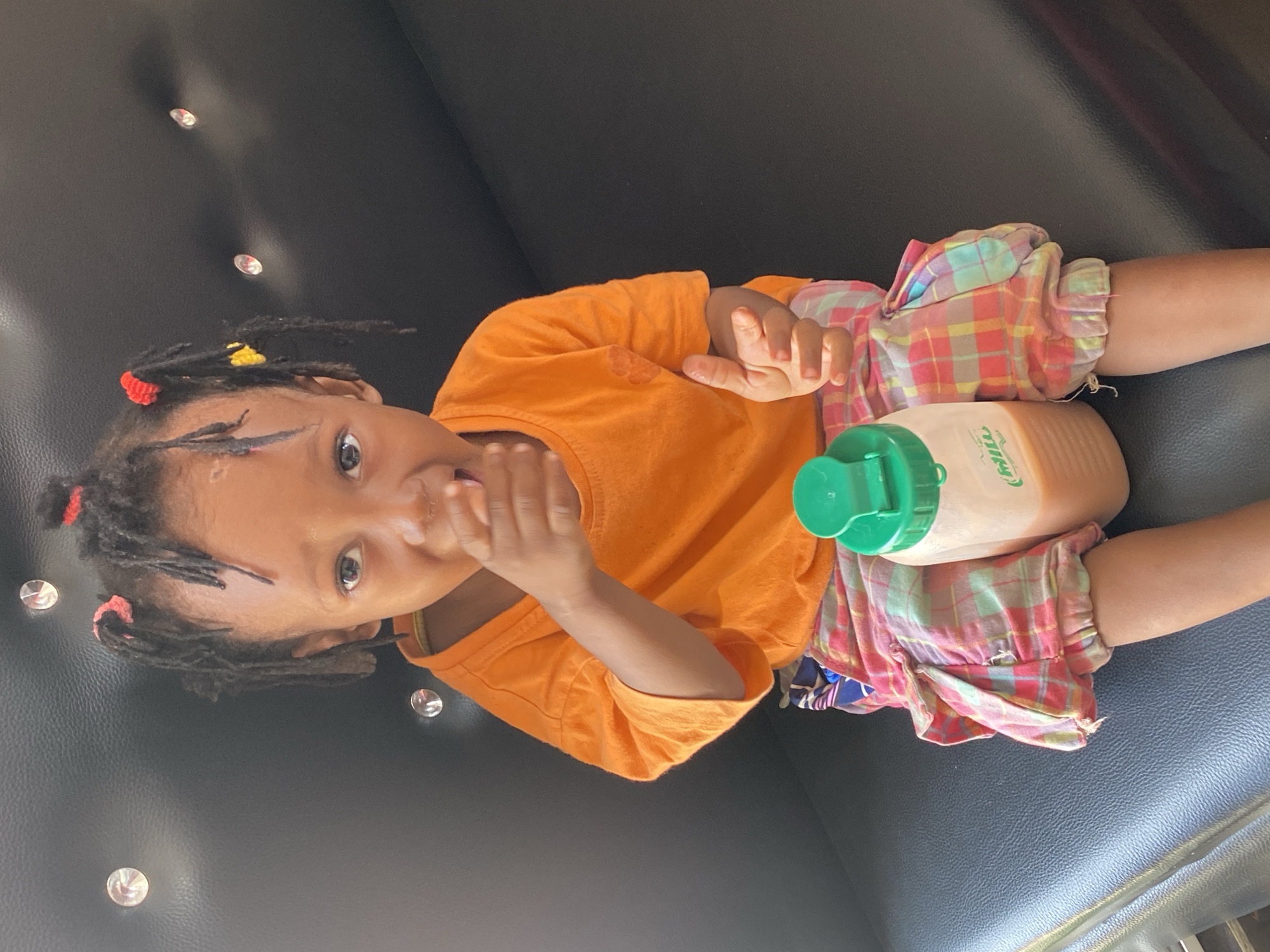 A young child is captured in a close-up, rotated image, lying on what appears to be a dark, padded surface, possibly a sofa or car seat, adorned with several small, reflective, studded embellishments. The child has dark skin and hair styled in numerous short dreadlocks or braids, accented with small red and yellow hair ties/clips. They are wearing a bright orange short-sleeved t-shirt and colorful plaid shorts featuring shades of pink, blue, yellow, and white.
The child is looking directly at the camera with wide, expressive eyes, and one hand is positioned near their mouth. Next to their left leg, a clear plastic bottle with a bright green lid rests, containing a light brown or beige liquid. The brand name "MILO" is visibly printed on the side of the bottle. The child's posture suggests they are relaxed, perhaps having a snack or drink. The setting appears to be indoors or within a vehicle. The child's expression is one of curiosity or engagement with the camera.