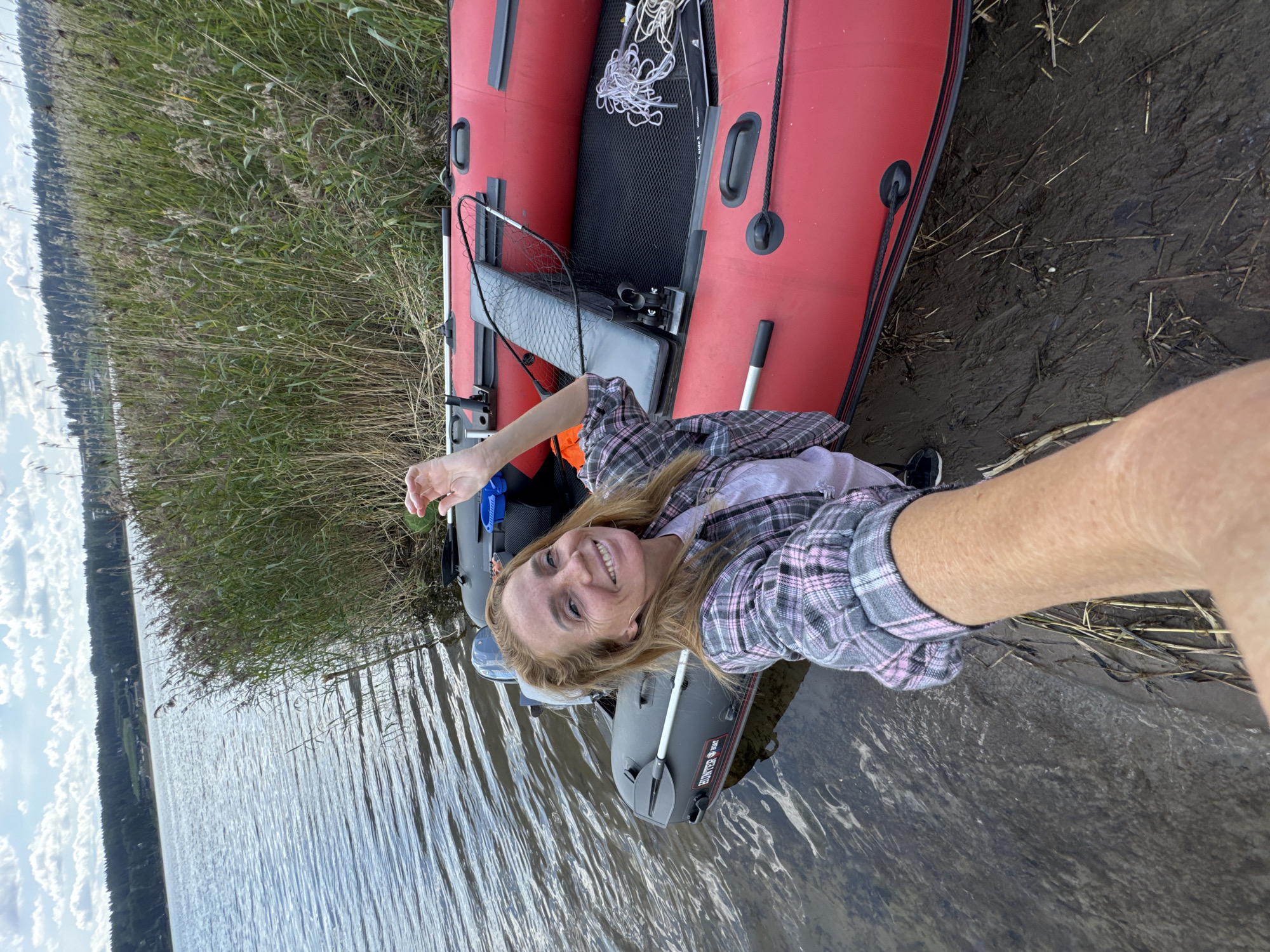 The image depicts a woman taking a selfie while lying on her back in a red inflatable boat, which is partially on muddy shore and partially in the water. She is smiling and looking at the camera, her right arm outstretched towards it. She is wearing a plaid shirt. The boat contains fishing gear, including a net and rope. The background shows a body of water with tall reeds along the shore, and a forested area in the distance. The sky is partly cloudy. The scene appears to be set during the day, with overcast weather. The location is identified as Portovoe, Russia. There is text visible on the grey inflatable boat in the foreground which says "HUNTER TRAIL".