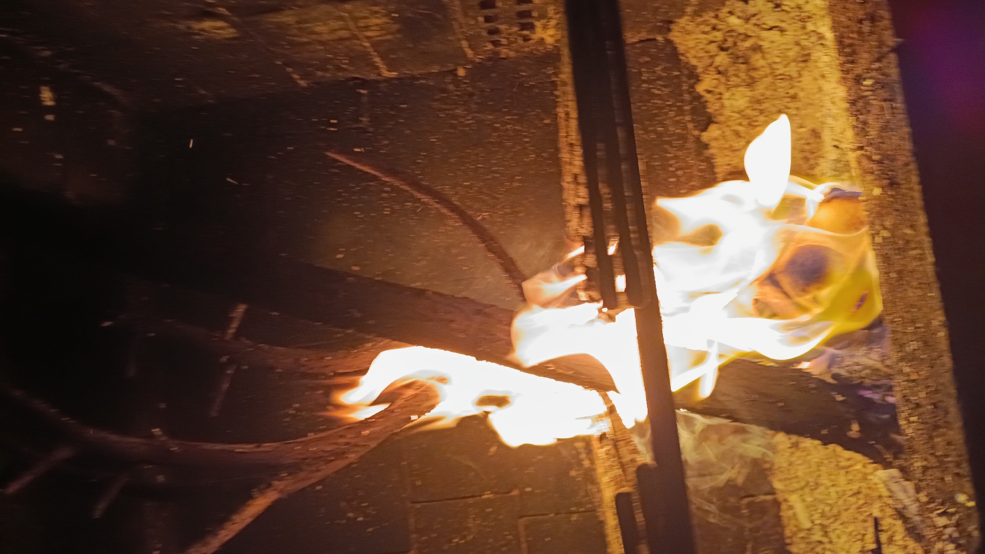 A fire burns in a brick fireplace. The flames are bright white and yellow, with orange and red hues at the edges. The fire is contained within a metal grate, and there are several pieces of wood burning. The background is dark and out of focus, but appears to be made of brick. The overall impression is one of warmth and intensity. This scene is located in the city of Novogusel'skii, Russia.