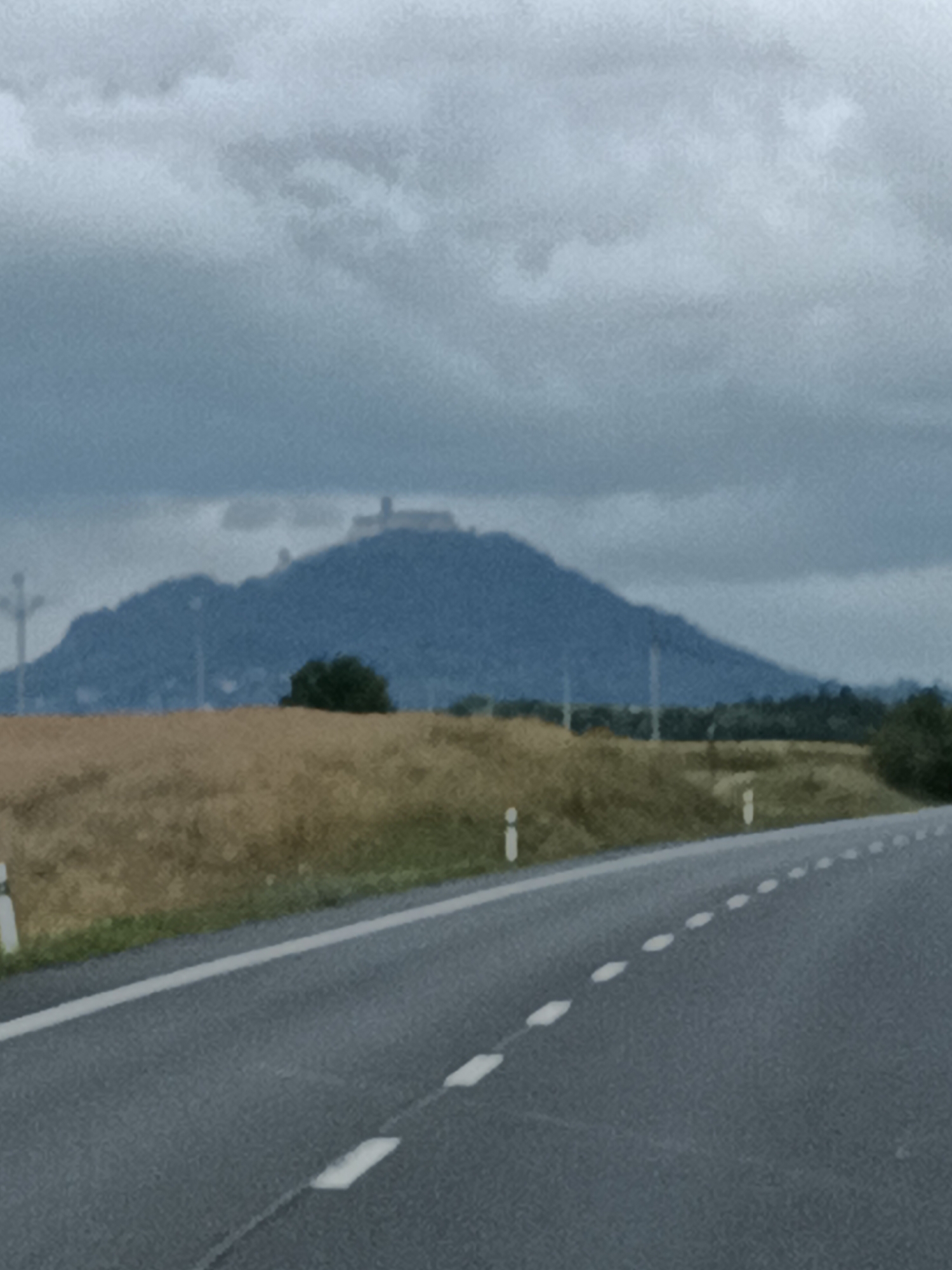 The image depicts a landscape scene with a road in the foreground, a field of dry grass in the middle ground, and a large hill with a structure on top in the background. The sky is overcast with clouds. The road has white dashed lines indicating lanes. The hill appears to be a prominent natural feature with a man-made structure, possibly a castle or observation point, situated on its summit. The vegetation in the middle ground is predominantly dry and appears to be a field of crops or grassland. There are a few scattered trees and some utility poles visible. The overall impression is one of a rural or semi-rural setting under a cloudy sky. The presence of a road suggests it is likely a view from a vehicle traveling along a highway or main road.