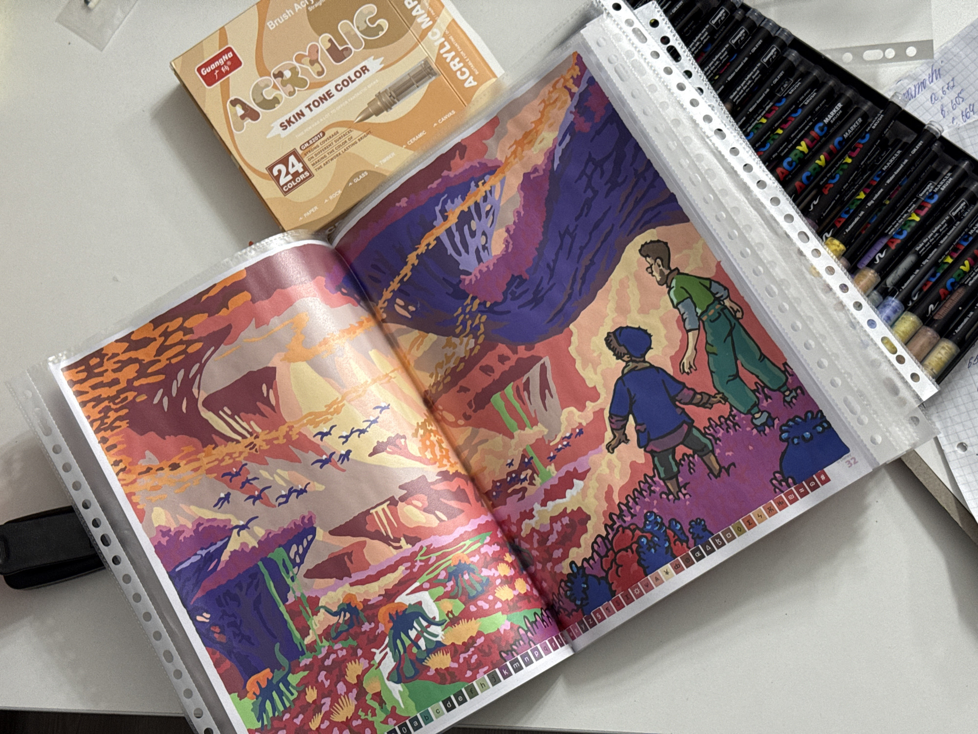 The image depicts a top-down view of a desk surface with art supplies and a print of a colorful illustration. In the foreground, a plastic binder holds two sheets of the illustration, which features a vibrant landscape with abstract elements and two figures in the lower section. The illustration uses a palette of reds, oranges, purples, blues, and greens. To the right of the binder, a box labeled "Guangxia Acrylic SKIN TONE COLOR 24 COLORS" is visible. Below the binder, a collection of acrylic markers with black barrels and colorful caps are arranged. A small notebook with handwritten notes is also present. The overall environment suggests an art workspace. There is no discernible location in Łódź, Poland.