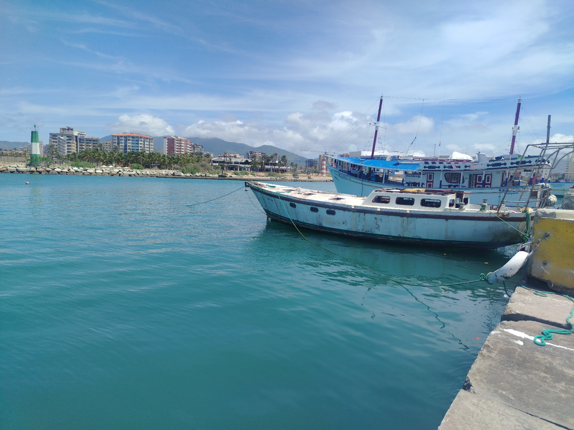 In Porlamar, Venezuela, a tranquil harbor scene unfolds under a bright, partly cloudy sky. The foreground is dominated by the calm, turquoise waters of the sea, reflecting the sunlight. Moored to a concrete dock, visible in the lower right quadrant, are two boats. The closer boat, a weathered, light-blue vessel, is tied with green ropes to a yellow fender attached to the dock. Its hull shows signs of age, with paint peeling and a visible waterline. Behind it, a larger, white boat with a blue canopy and red accents is also docked. Its masts and rigging are prominent against the sky. In the background, the cityscape of Porlamar stretches along the coastline, characterized by a cluster of multi-story buildings and a lighthouse with green and white bands. Palm trees line the shore in front of the buildings, and lush green mountains rise in the distance, completing the scenic backdrop of this coastal Venezuelan city.