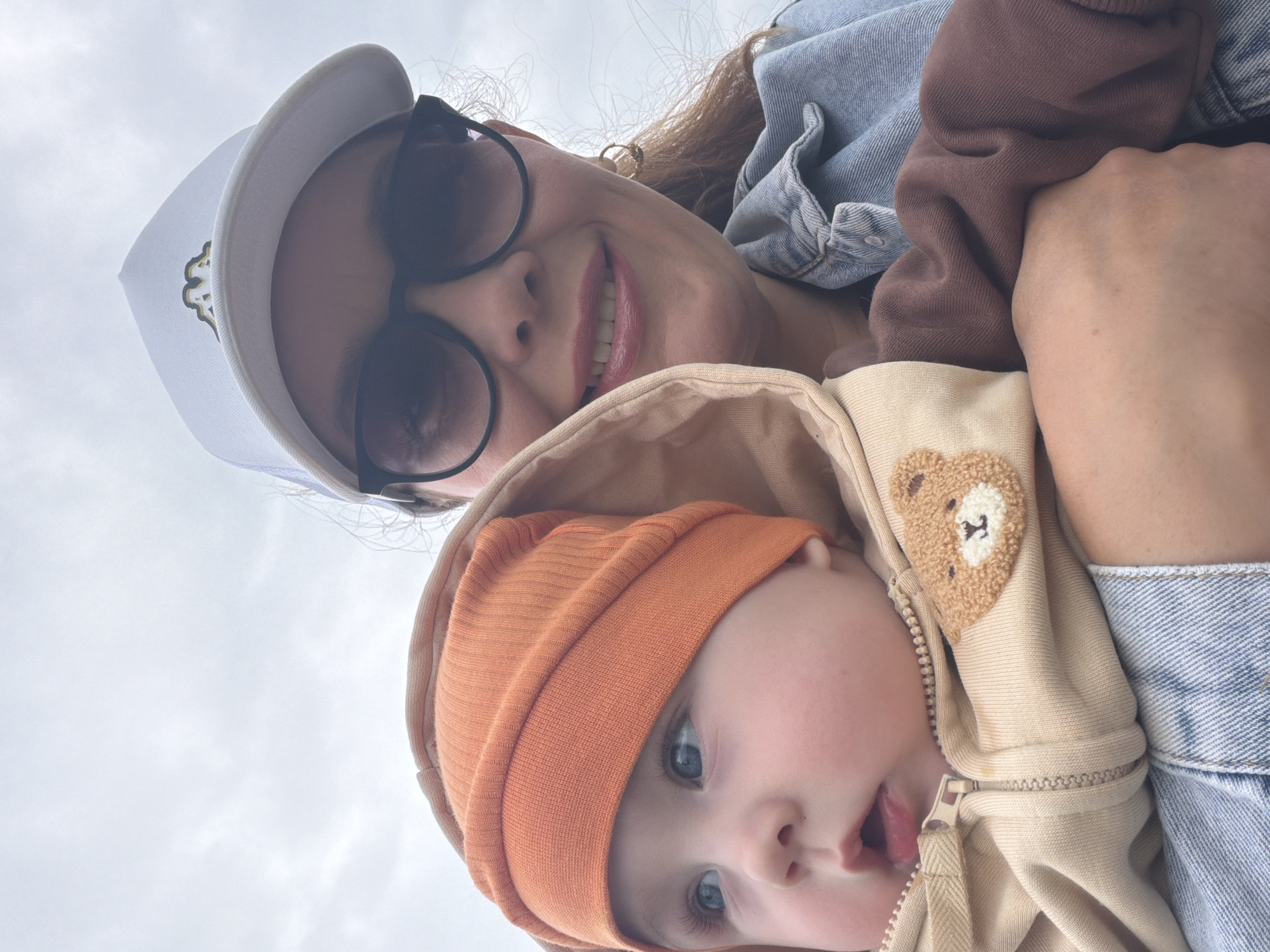 An outdoor, medium shot photograph depicts a woman holding a baby. The woman is wearing a white baseball cap with a gold logo, dark sunglasses, and a denim jacket. She is smiling and looking at the camera. The baby is wearing an orange beanie and a tan hooded jacket with a teddy bear appliqué on the chest. The baby is looking to the left of the frame. The background is a cloudy, overcast sky, suggesting an outdoor location in Chaikovsky, Russia, likely during daylight hours. The overall impression is one of a casual, affectionate moment between a mother and child.