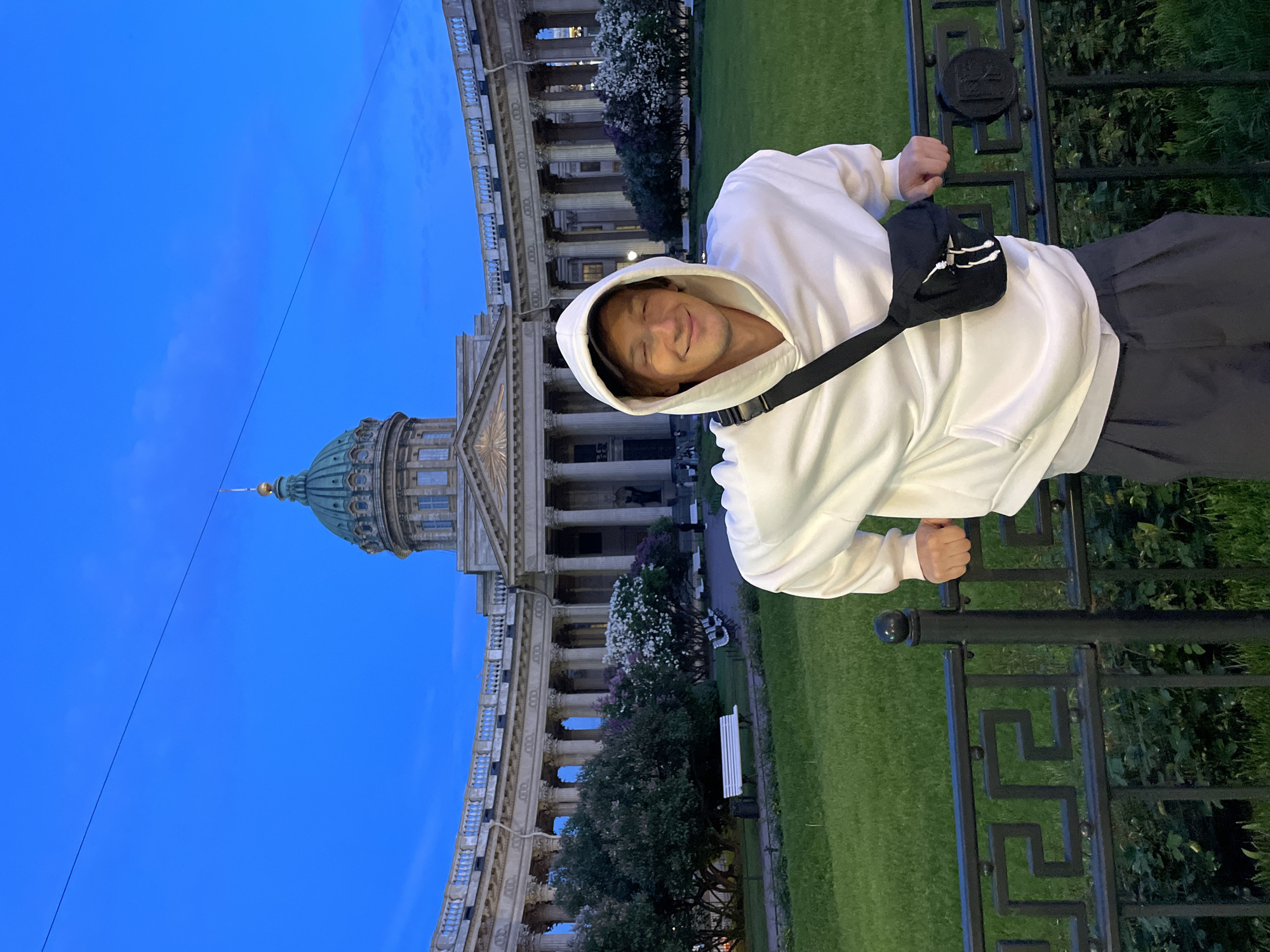 The image displays a young man posing in front of a grand architectural landmark, likely the Kazan Cathedral in Saint Petersburg, Russia. The scene unfolds outdoors during the "blue hour," characterized by a clear, deep blue sky that occupies a significant portion of the background. A thin black wire stretches horizontally across the sky.
The subject is a young man, smiling broadly with his eyes squinted, suggesting a cheerful or relaxed mood. He has light brown hair and is wearing a white hooded sweatshirt, dark pants, and a black crossbody bag with three white stripes. He is leaning casually against a decorative black metal fence, grasping the top rail with both hands. The fence features intricate geometric patterns, and a circular emblem resembling a "no smoking" sign (a cigarette with a diagonal line through it) is visible on it.
Behind the man, the Kazan Cathedral stands majestically. It is a large, classical building with numerous tall columns forming a colonnade, a distinctive green dome topped with a golden spire, and a balustrade. In front of the cathedral, a well-maintained green lawn is visible, along with lush trees and bushes, some adorned with white and purple blossoms. A white park bench can be partially seen among the greenery. The overall setting depicts a serene public square or park, with the historical architecture serving as an imposing backdrop.