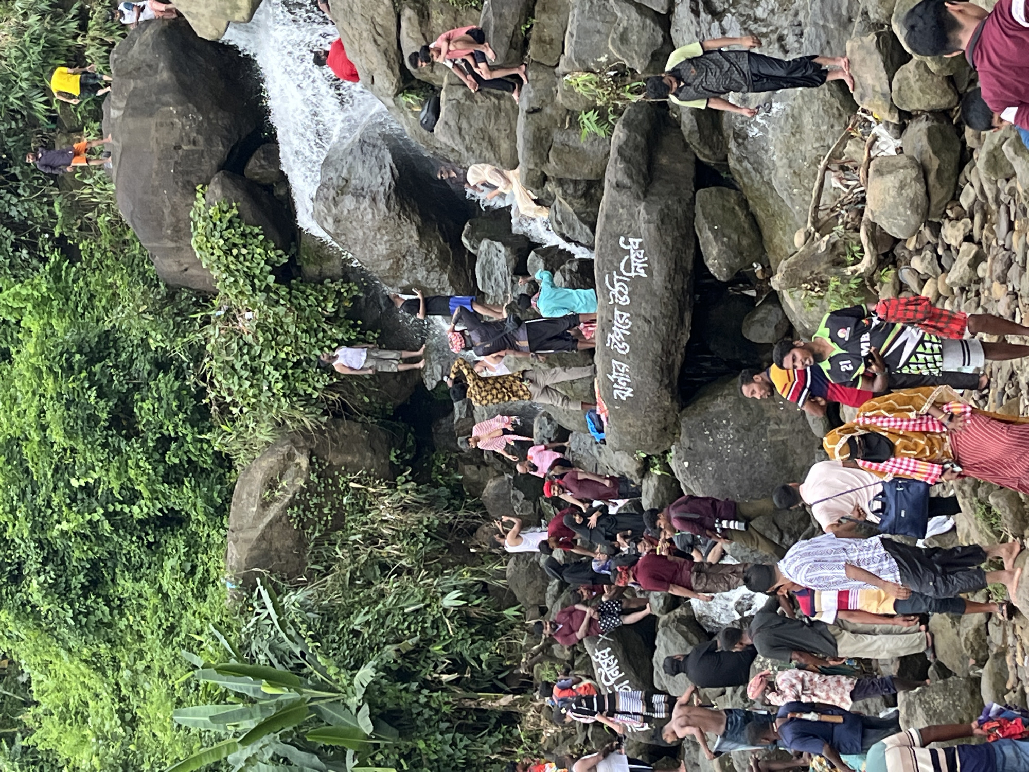 The image depicts a lively scene at a waterfall in Jaflong, Bangladesh. A diverse group of people are scattered across rocks and near the cascading water, engaged in various activities. Some individuals are standing on the rocks, observing the scenery, while others are seated. A few people appear to be wading in the water, enjoying the cool spray. Several individuals are dressed in casual clothing, including t-shirts, shorts, and traditional attire. Among the crowd, one person is seen taking a photograph, suggesting the scenic beauty of the location. The environment is characterized by lush green vegetation covering the rocky terrain and the presence of a significant waterfall. The atmosphere appears to be one of leisure and enjoyment, with many people gathered to experience the natural attraction.