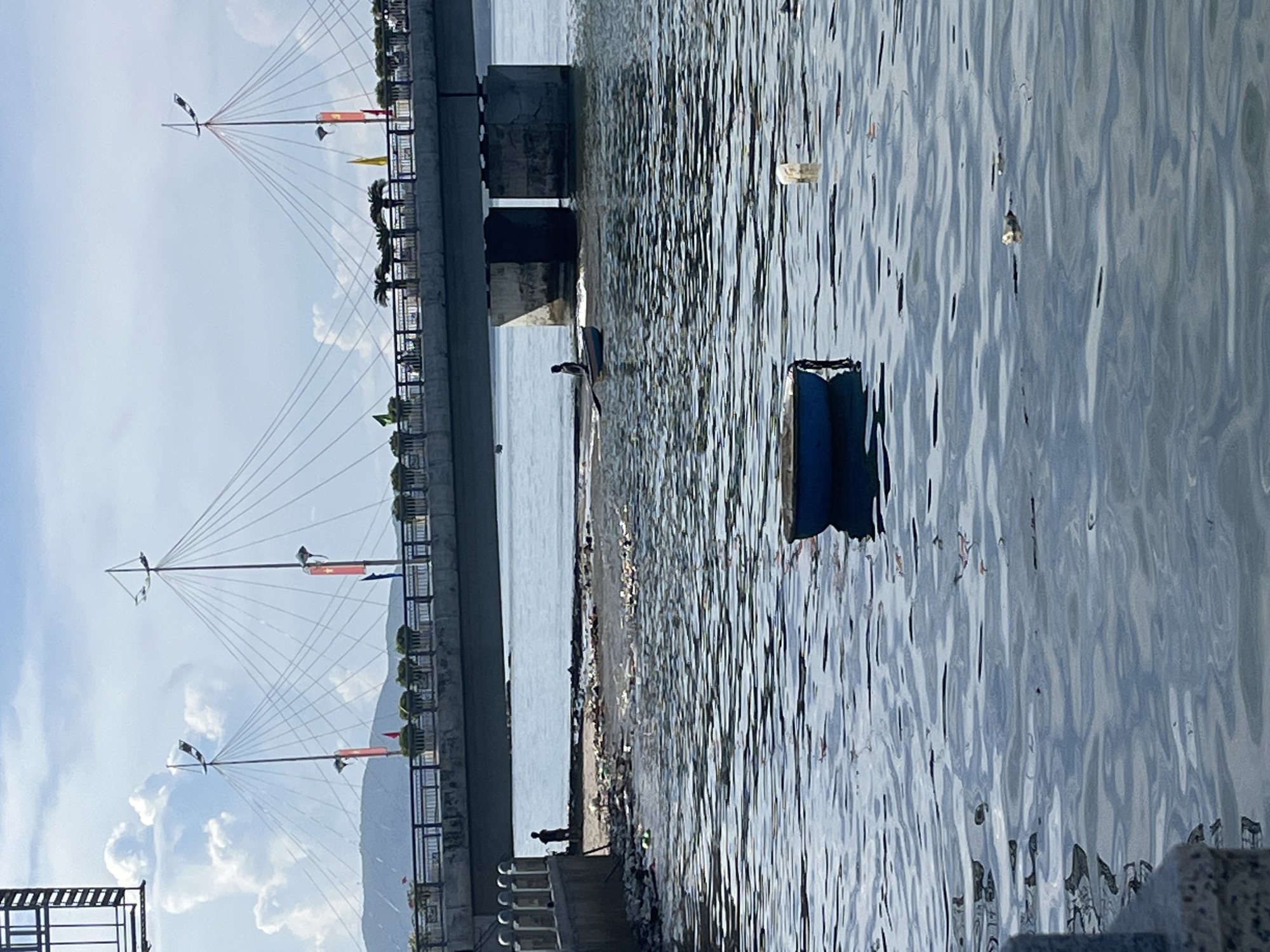 The image depicts a view of the sea in Bac Nha Trang Ward, Vietnam, with a bridge structure visible on the left. The bridge features decorative towers with multiple cables extending from their apexes. The water surface is rippled, with debris floating in it. The bridge supports are visible, with what appears to be a person standing near one of the supports. The shoreline along the bridge is littered with debris. In the background, there's a glimpse of a mountainous landscape and cloudy sky.