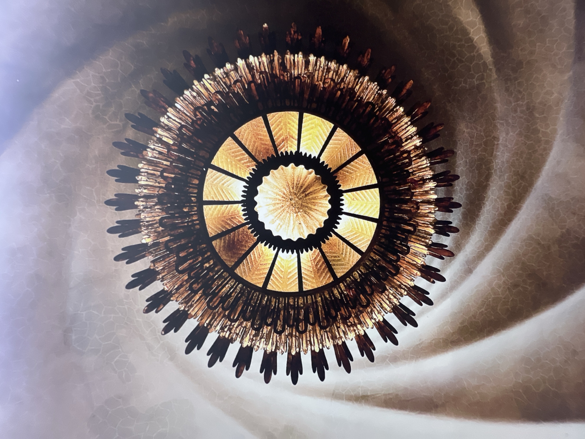 This is a close-up, upward-facing shot of an ornate, starburst-shaped chandelier hanging from a ceiling. The chandelier is primarily made of dark, metallic rods and thin, pointed, amber-colored glass elements that radiate outwards from a central, glowing light source. The light creates a warm, golden illumination and casts shadows. The ceiling has a textured, swirling pattern, appearing to be made of a light-colored material with darker, irregular markings. The overall impression is one of intricate design and soft, ambient lighting. This chandelier is located in the Casa Batlló, Barcelona, Spain.