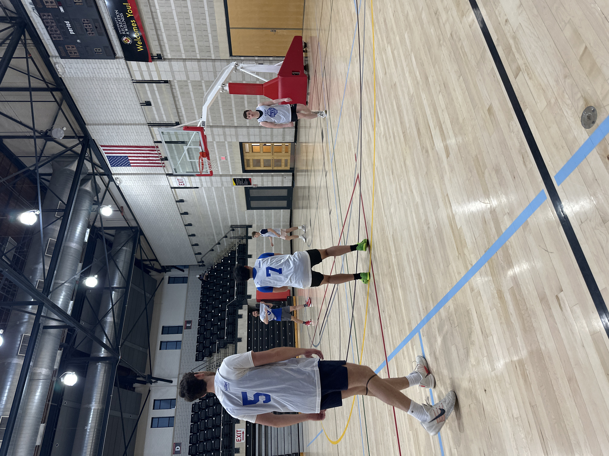 The image captures a basketball court inside a gymnasium at the University of Maryland in College Park, United States of America. Several individuals wearing basketball jerseys are visible, each adorned with a number. The closest person wears jersey number 5 with "Hillel" and the second person wears jersey number 7 with "Hillel." They are standing on a light-colored wooden floor marked with various colored lines indicating the court boundaries. Above the court, one basketball hoop is visible with a red backboard, and in the background, there are bleacher seats. An American flag hangs above a doorway marked "EXIT," and a scoreboard displaying the word "PERIOD" is above this. A banner reading "University Recreation & Wellness Welcomes You!" hangs on the wall. The structure of the gymnasium includes metal beams and ventilation ducts visible on the ceiling.