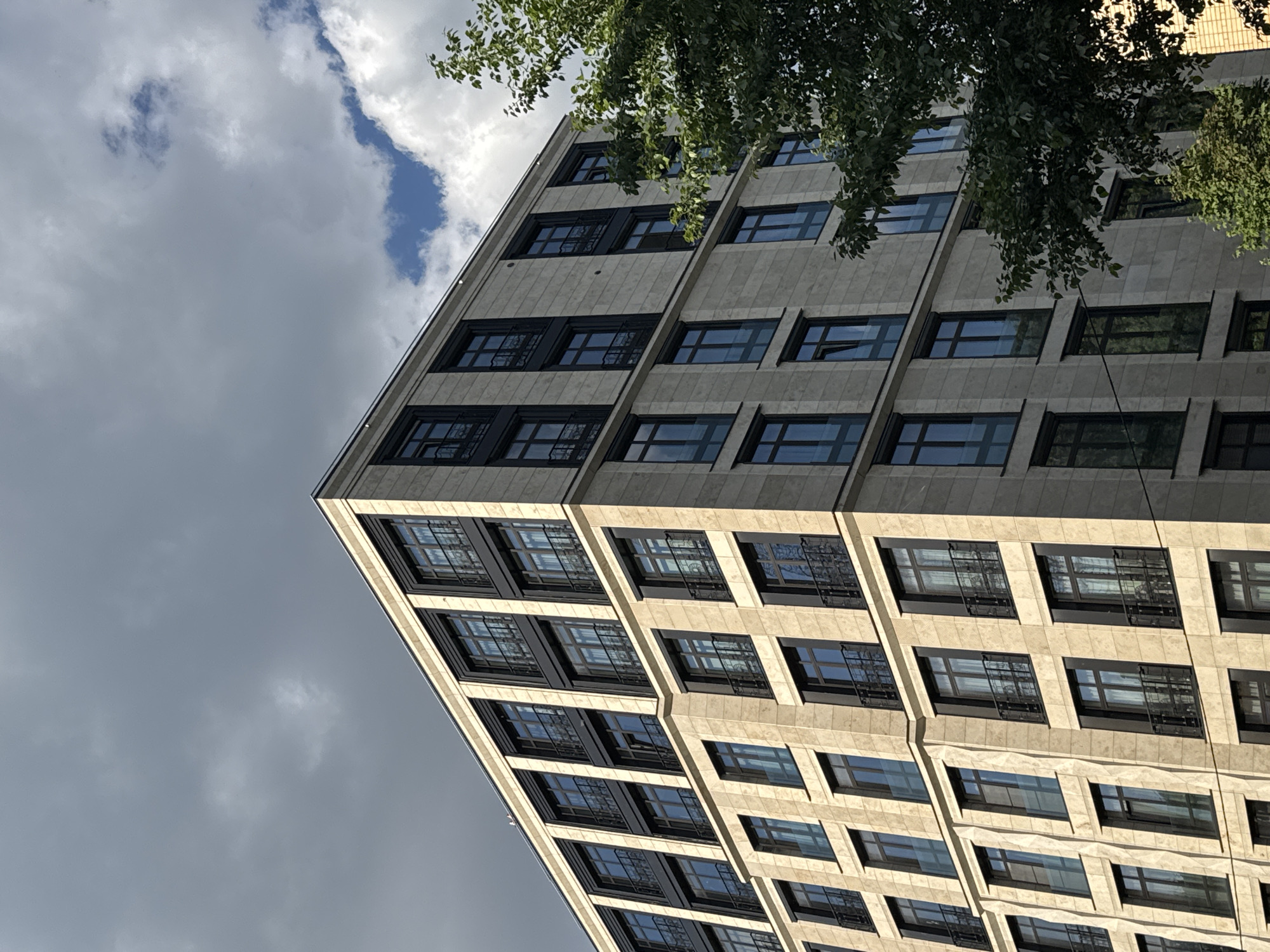 The image captures a modern, multi-story building from a low-angle perspective, looking upwards towards the sky. The building's facade is a light, possibly beige or off-white, textured material, structured with a grid of recessed, dark-framed windows. These windows appear dark or show subtle reflections of the sky, and many have horizontal mullions. The building extends diagonally from the lower left towards the upper right of the frame, showcasing its angular architecture and multiple floors.
In the upper right corner, a significant portion of dark green foliage from a tree hangs over the building, partially obscuring the upper floors and adding a natural element to the urban scene. The sky, visible in the upper left and central areas, is partially cloudy, featuring patches of bright blue sky interspersed with light and dark grey clouds. Sunlight illuminates parts of the building's facade, suggesting it is daytime. There are no people visible, nor any discernible text. The scene is an objective architectural shot within an urban setting.