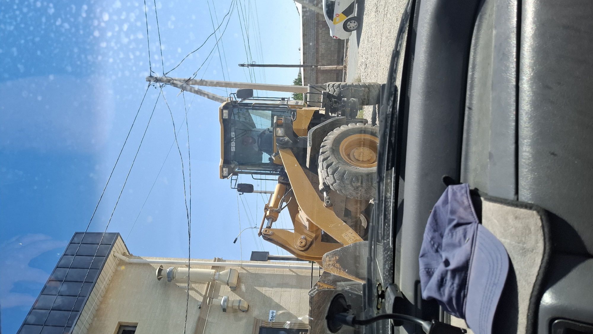 In Шұбарсу, Kazakhstan, a yellow front-end loader is parked on a gravel surface next to a building. The loader has a bucket extended forward and a person is visible in the driver's seat. A utility pole with wires stands nearby. In the foreground, a blue baseball cap rests on a car's dashboard. A white car with signage is visible in the background. The sky is clear and blue.