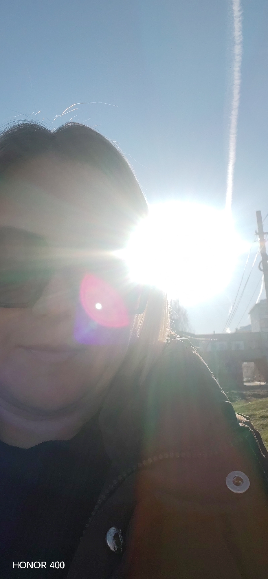 A selfie taken outdoors under bright sunlight obscuring most of the face of a person wearing dark glasses and a dark jacket with metal fasteners. A contrail is visible in the sky. The background includes a building, a structure resembling a bridge, and utility poles with wires. The phone brand "HONOR 400" is visible in the lower left corner. The specific location in the city of Noviy is not clear enough to tell from these objects.