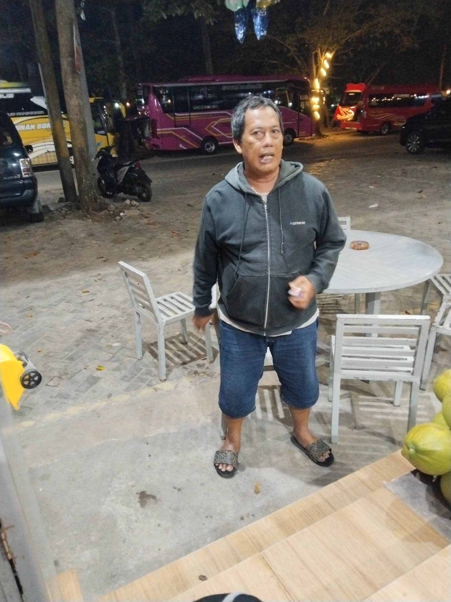 A middle-aged man stands in front of a round table with chairs, appearing to be at an outdoor seating area near a road. He is wearing a dark grey hooded jacket with the word "converse" on it, blue denim shorts, and patterned sandals. The man is looking towards the camera with his mouth slightly open. In the background, several buses are parked. One is pink with white stripes, another is red, and there is a yellow bus partially visible. A black motorcycle is parked to the left of the pink bus. The ground is a mix of cobblestones and concrete. The lighting suggests it is nighttime or dusk, with artificial lights visible in the trees behind the buses. The overall scene appears to be a rest stop or a parking area in Prigi, Indonesia.