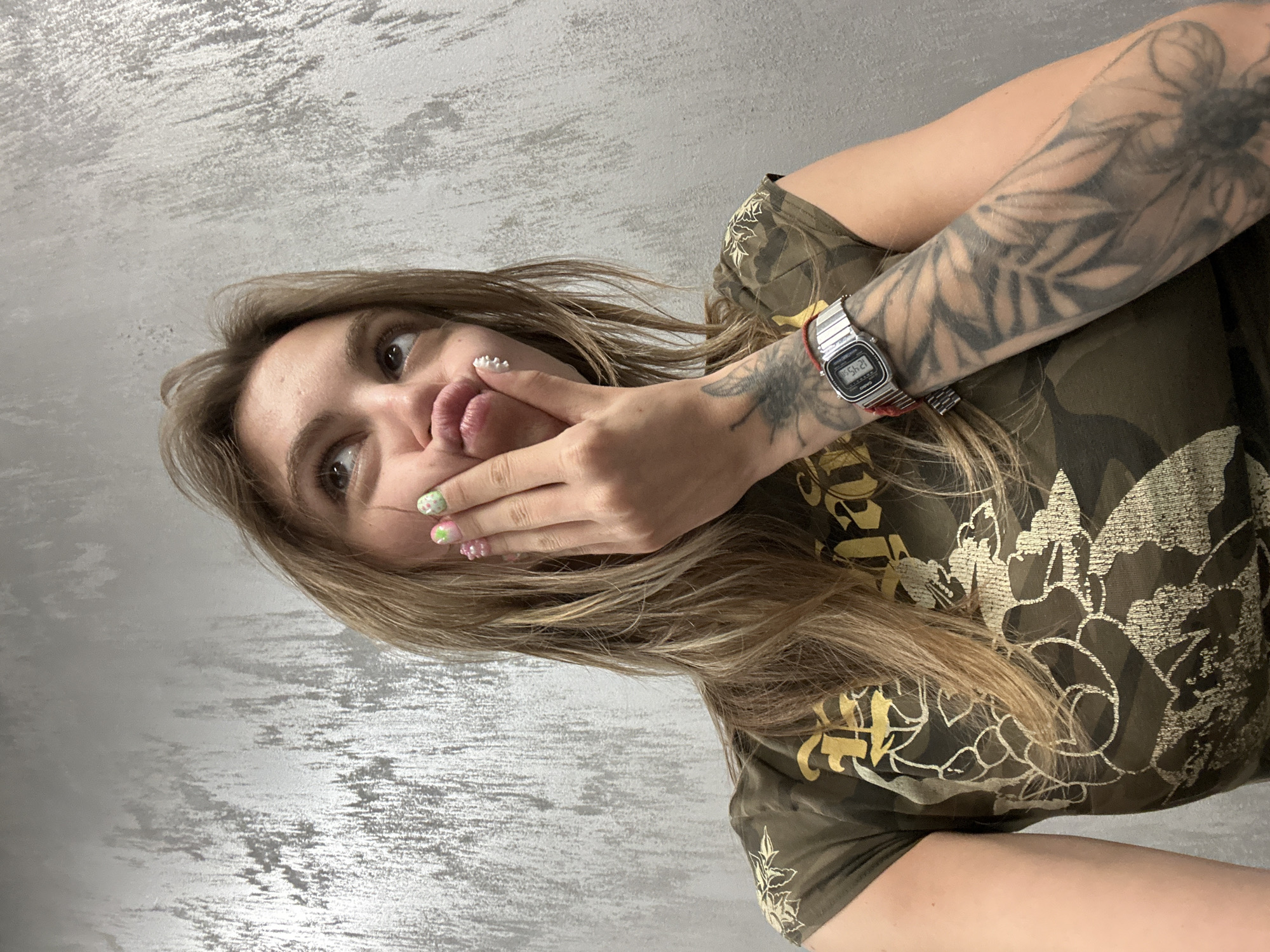 A medium shot captures a young woman with long blonde hair, wearing a camouflage t-shirt with gold floral patterns. She is holding her right hand to her chin, with her lips puckered and eyes looking upward. Her fingernails are decorated with colorful, playful designs, including what appear to be small charms or beads. A silver watch with a red band is visible on her left wrist. The background is a textured gray wall. The image provides no contextual information about its location in Moscow, Russia, or any specific details about the environment beyond the visible wall.