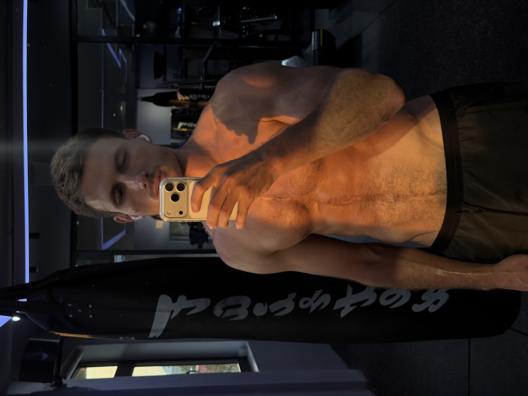 A shirtless man is taking a selfie in a gym or fitness area. He is fair-skinned with short hair and appears to be in good physical condition. He is holding a smartphone to take the picture, and a pair of wireless earbuds is visible in his ear. Behind him, there is a black punching bag with white lettering on it. The background reveals gym equipment and mirrored surfaces, suggesting the location is a fitness facility. There is no specific indication that it is in Đà Nẵng, Vietnam.
