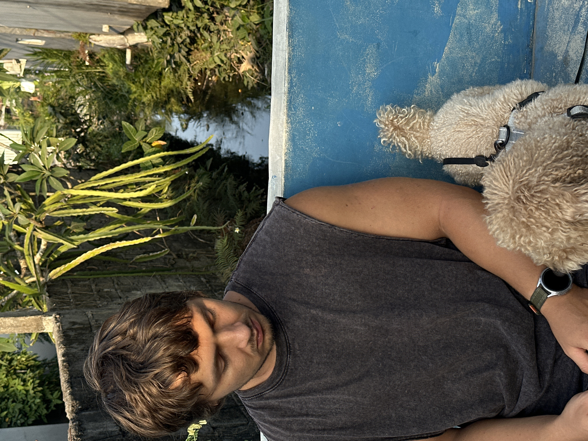 A young man with short brown hair and a gray sleeveless shirt sits on a blue bench, looking down. Next to him, a small, light-colored dog with curly fur faces away from the camera, wearing a black harness. The man has a wristwatch on his left wrist. Behind them, a narrow waterway is visible, bordered by lush green vegetation, including hanging, elongated green plant pods, and ferns. A rough, gray stone wall is to the left, and a wooden structure is on the right. The scene appears to be outdoors, possibly in a garden or near a canal in North Kuta, Indonesia.