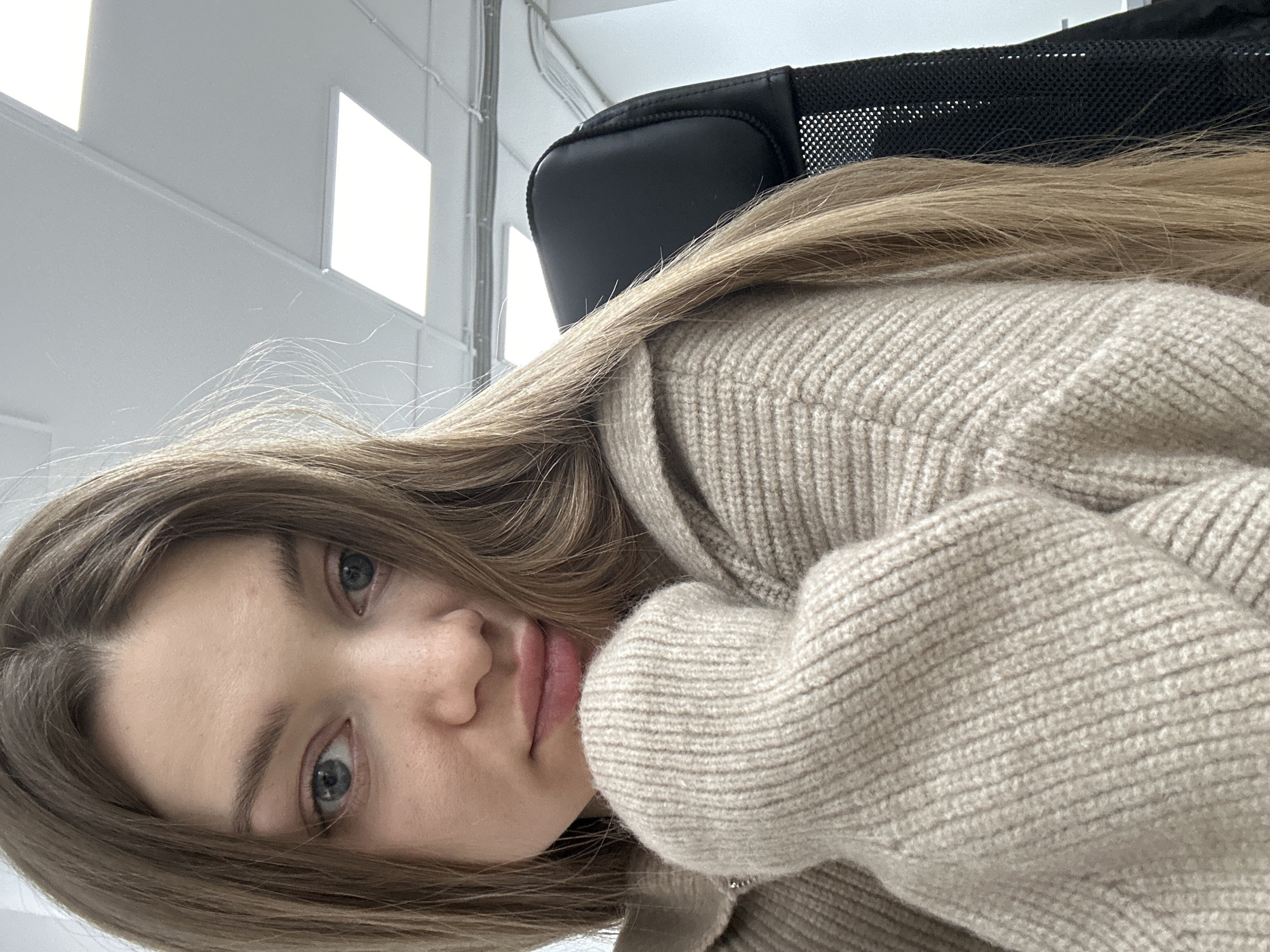 A selfie shows a fair-skinned young woman with light brown hair and blue eyes. She wears a beige knit sweater. The background shows an office setting, with overhead lights visible near the top and a black office chair behind her. There is no verifiable information indicating this was taken in Kazan, Russia.
