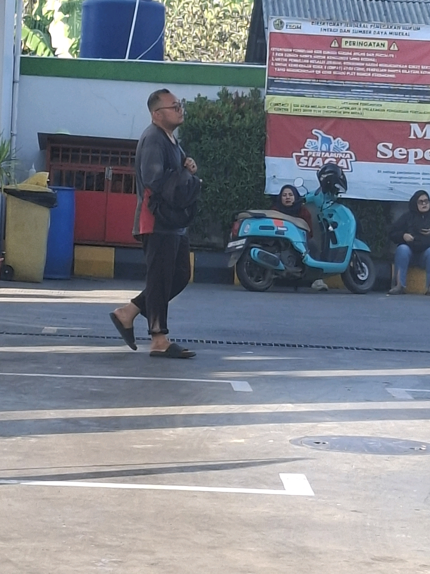A man in a dark gray long-sleeved shirt and dark pants is walking on an asphalt surface, wearing dark flip-flops. He has glasses and is looking upwards. In the background, there is a light blue motorcycle with two people on it, a woman in a dark headscarf and a dark jacket, and another person behind her wearing glasses and a dark jacket. To the left of the motorcycle, a yellow trash bin is visible. Behind the motorcycle, a large red and white sign with Indonesian text and the Pertamina logo is displayed. The environment appears to be outdoors in a city setting, possibly a street or parking area, with a building structure and vegetation in the background. The overall lighting suggests it is daytime.