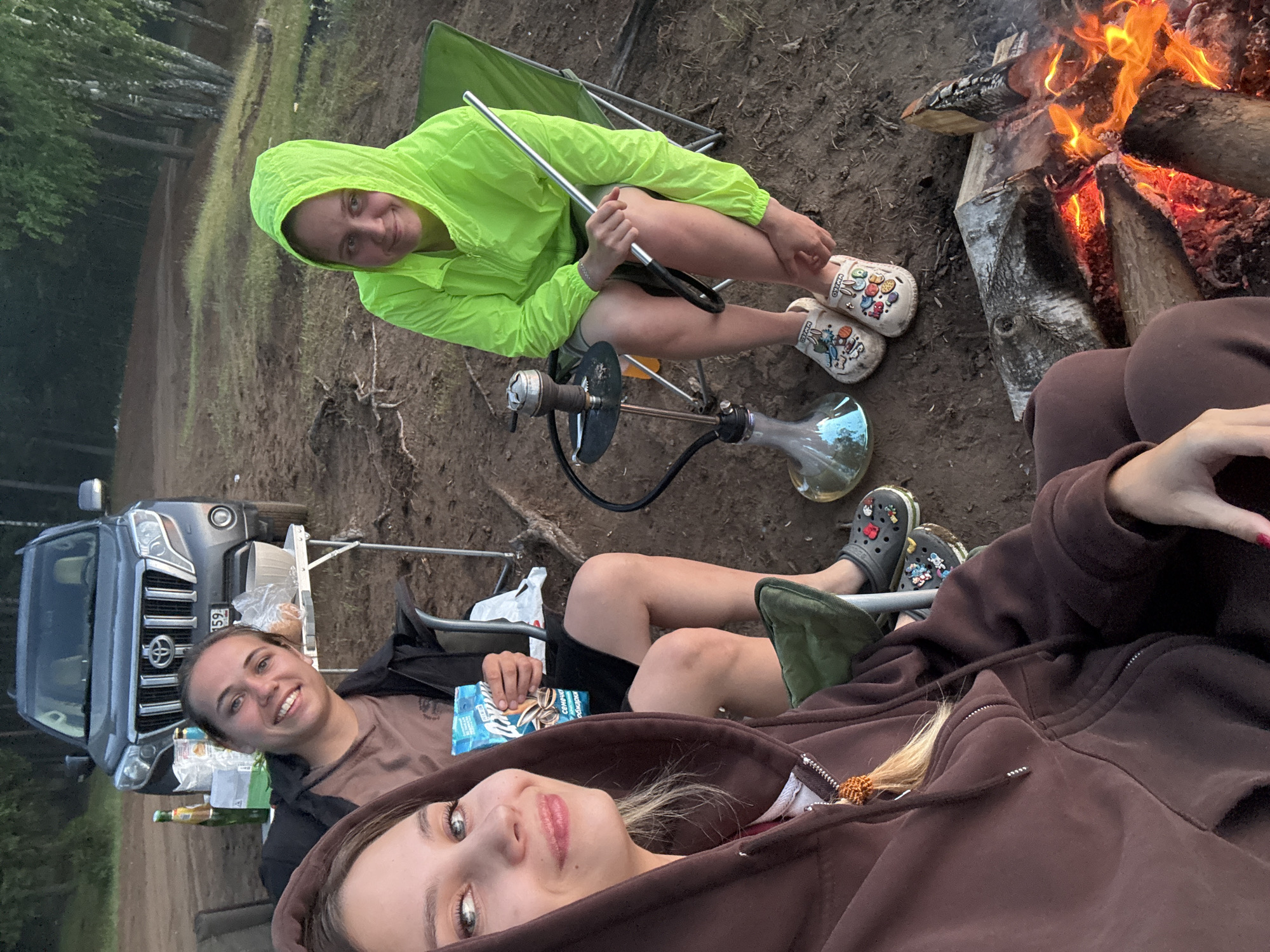 In the city of Chaikovsky, Russia, three young women are gathered around a campfire. Two women are seated in folding chairs, and a third is in the foreground, partially visible. A dark gray Toyota Land Cruiser Prado is parked in the background. A portable table with food and drinks sits behind the two seated women. A hookah is set up on the ground, and a young woman in a neon green hooded jacket is holding its hose. Another woman, wearing a brown hooded sweatshirt, is holding a bag of sunflower seeds. A fire pit with burning logs is in the foreground. The setting appears to be a natural outdoor area, likely on the outskirts of the city, with trees and grassy terrain visible.