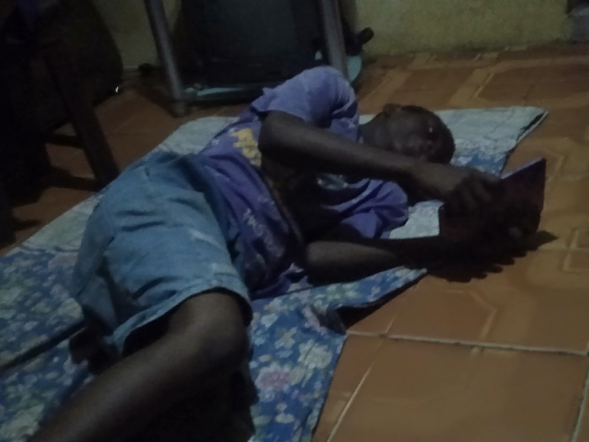 The scene depicts an individual lying on a patterned mat on a tiled floor. The person is wearing a purple t-shirt and blue shorts, and is holding what appears to be a book or tablet in their hands. The location appears to be indoors, likely a private residence. The specific city and region are unknown. A chair and other furniture are partially visible in the background. The individual appears to be reading or using the device.