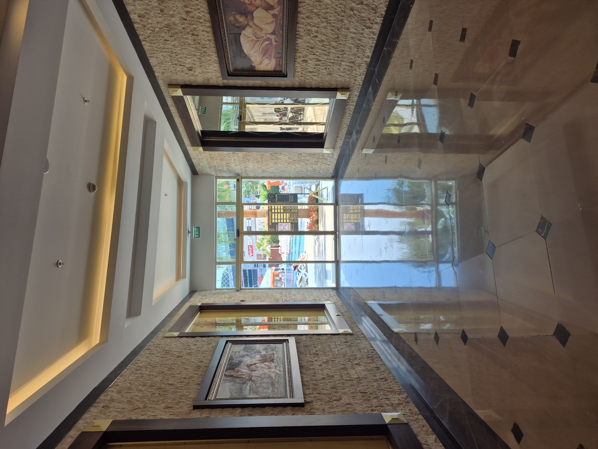 The image depicts the interior of a building, likely a hotel or resort, located in Alanya, Turkey. It is oriented looking towards a window with a view of the exterior. The floor is tiled with a dark border and has a reflective surface. The walls are covered with a textured stone material and feature framed artwork at varying heights. There are horizontal mirrors alternating between the artworks. The ceiling has recessed lighting. Outside the window is a view of a pool, palm trees, and a building with signs in Turkish. The exit sign near the window suggests this might be an emergency exit.
