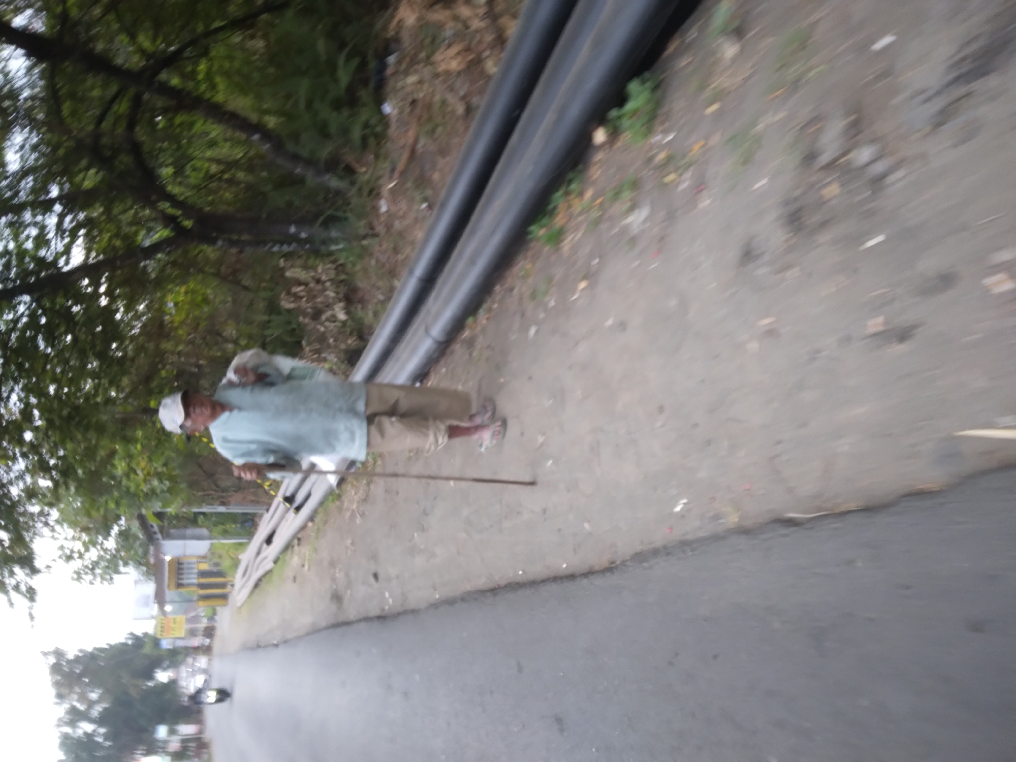 The image depicts an elderly man wearing a light blue shirt and beige pants, a white cap, and sandals, walking with a stick on a paved road. He is holding a white cloth draped over his left shoulder and the stick in his right hand. To the right of the road, there are several large black pipes lying on the ground, with some vegetation and debris around them. In the background, there are trees and some buildings. The scene is likely located in Desa Pamijen, Kecamatan Sokaraja, Indonesia, given the context provided. The overall atmosphere of the image is that of a common street scene in a rural or semi-urban Indonesian setting.