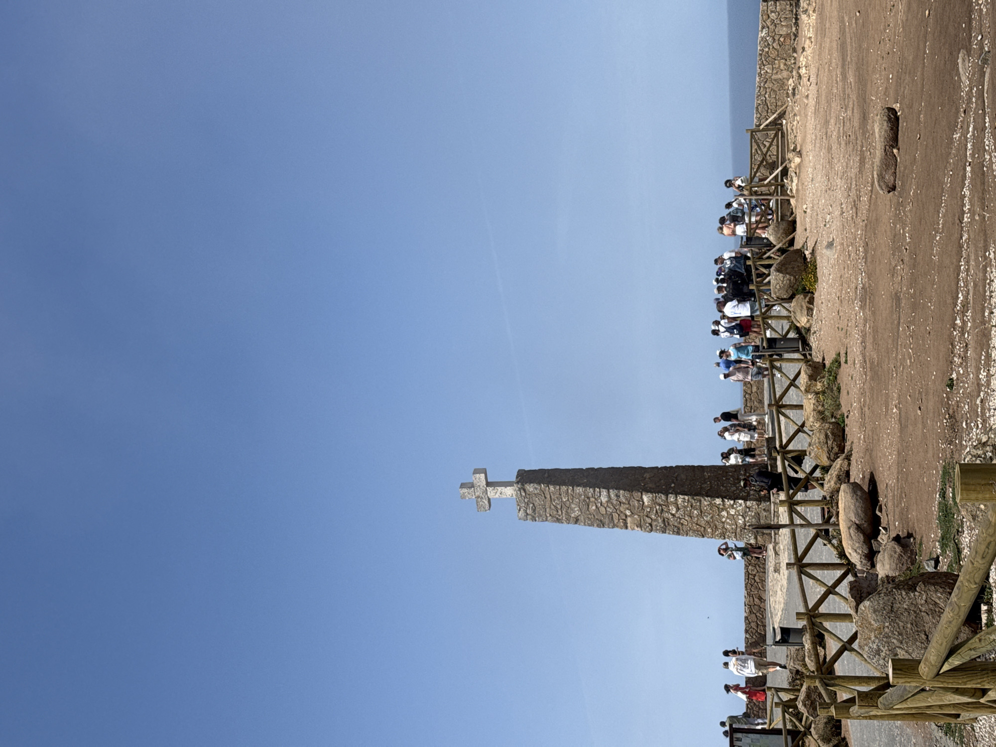 The image shows the monument at Cabo da Roca, the westernmost point of mainland Europe, located in the municipality of Colares, Portugal. A tall, stone monument topped with a cross stands prominently against a clear blue sky. A group of tourists is gathered around the monument, some standing on a paved area behind a wooden fence, and others further back on a stone wall overlooking the ocean. The ground in the foreground is a mix of dirt, rocks, and gravel, with more wooden fencing. The ocean is visible in the background. The scene depicts a popular tourist destination on a sunny day.