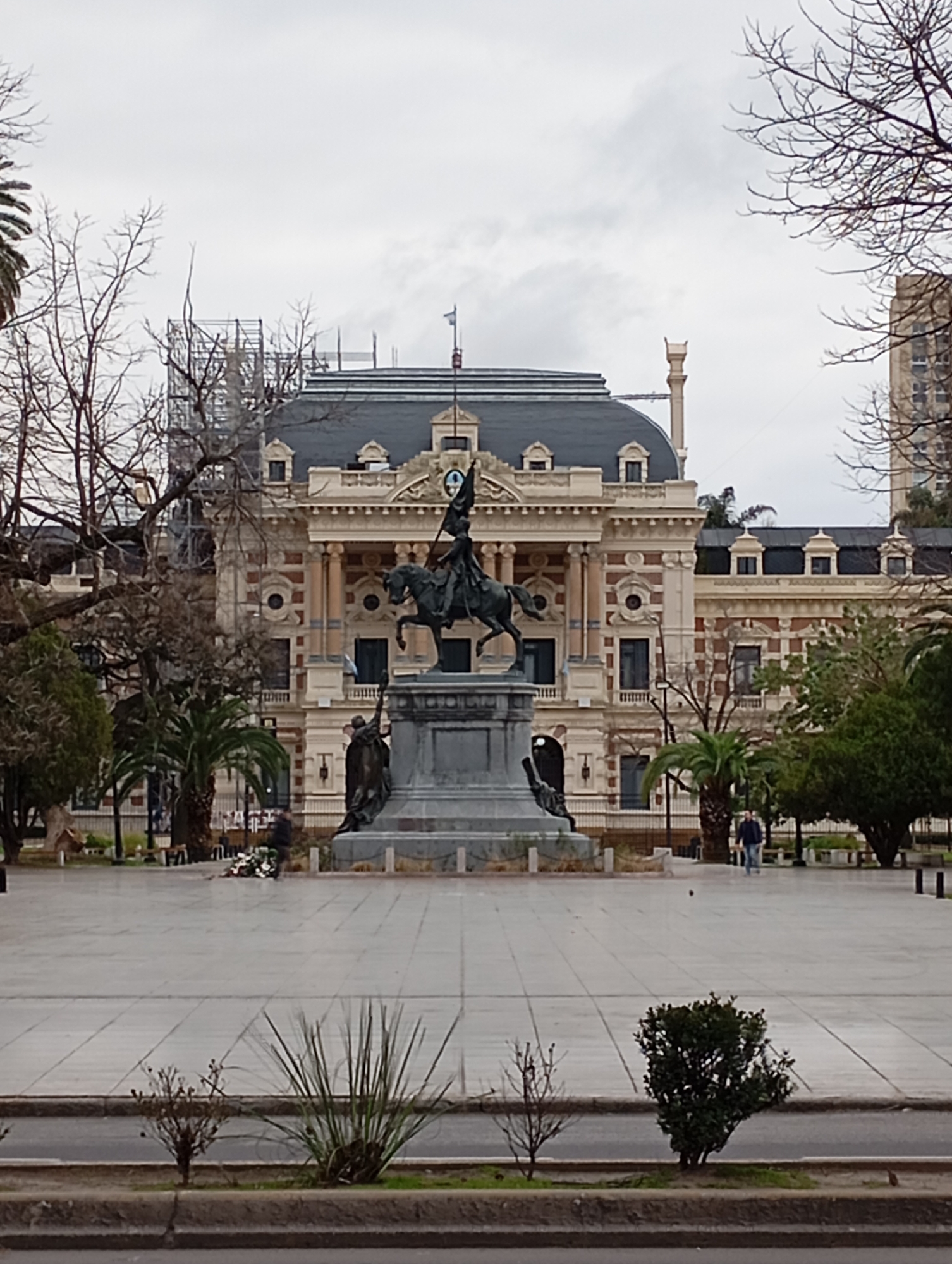 The image depicts the Plaza Moreno in the city of La Plata, Argentina, under an overcast sky. Dominating the central area is the Monumento a la Bandera, a large equestrian statue of a figure holding a flag, mounted on a substantial stone pedestal. At the base of the pedestal, two additional sculpted figures are visible, one with a draped garment. To the right of the monument, a person in a dark jacket and blue jeans walks away from the viewer. Further to the right, another person is seen standing. In the background, the impressive architecture of the La Plata Cathedral is partially obscured by trees and scaffolding, suggesting ongoing construction or maintenance. Palm trees and deciduous trees with bare branches are interspersed throughout the plaza. The ground is paved with large, light-colored tiles, creating a vast open space. In the foreground, along the edge of the paved area, there is a strip of grass with several small bushes and a larger, dense bush on the right.